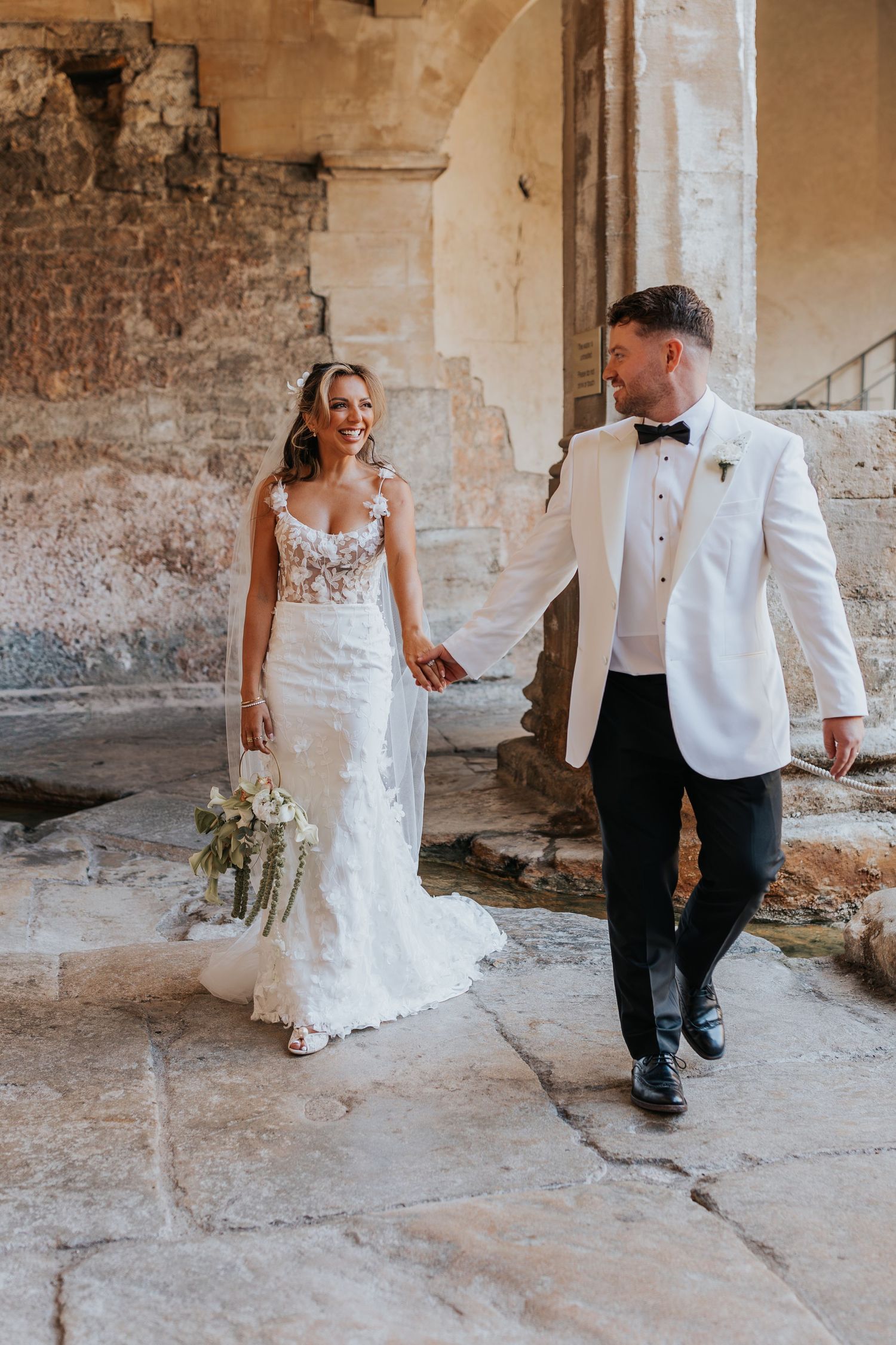 Bride and groom walk hand-in-hand at a rustic stone venue, she in a lace gown, he in a white tuxedo jacket.