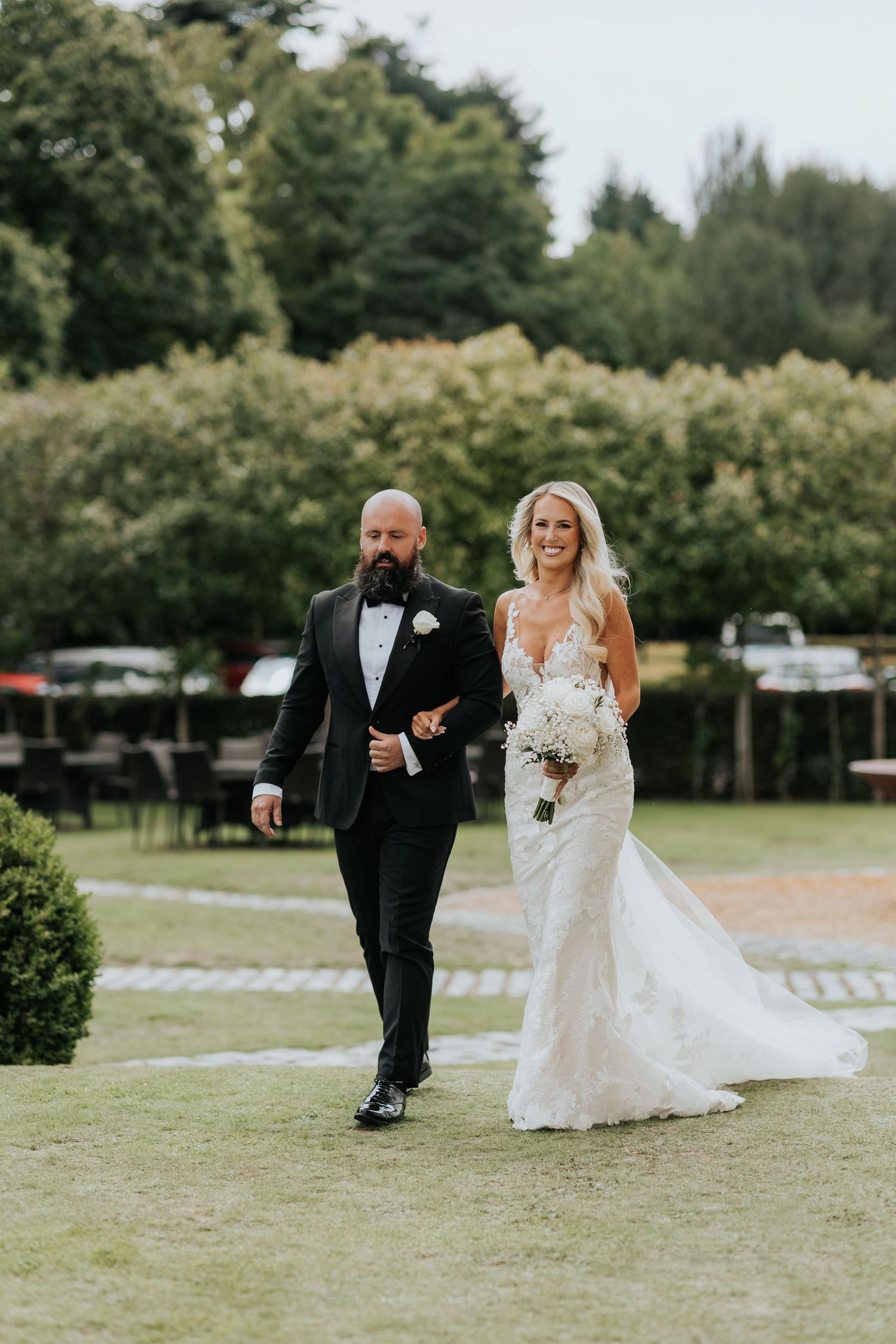 Bride in lace gown holding white bouquet walks with bearded man in black tuxedo on green lawn at outdoor wedding.