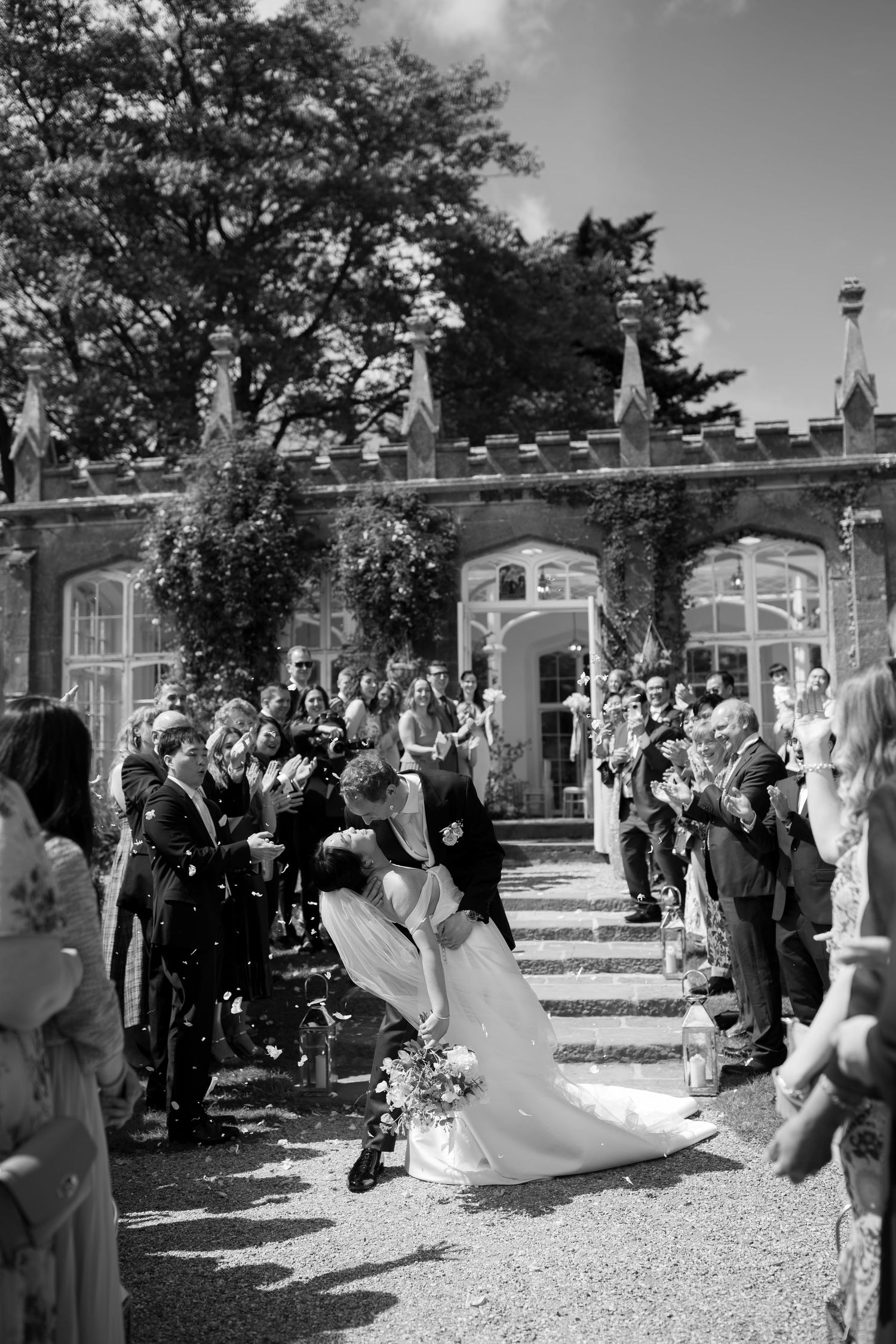 Black and white photo of a bride and groom sharing a kiss while guests throw confetti outside an elegant venue.