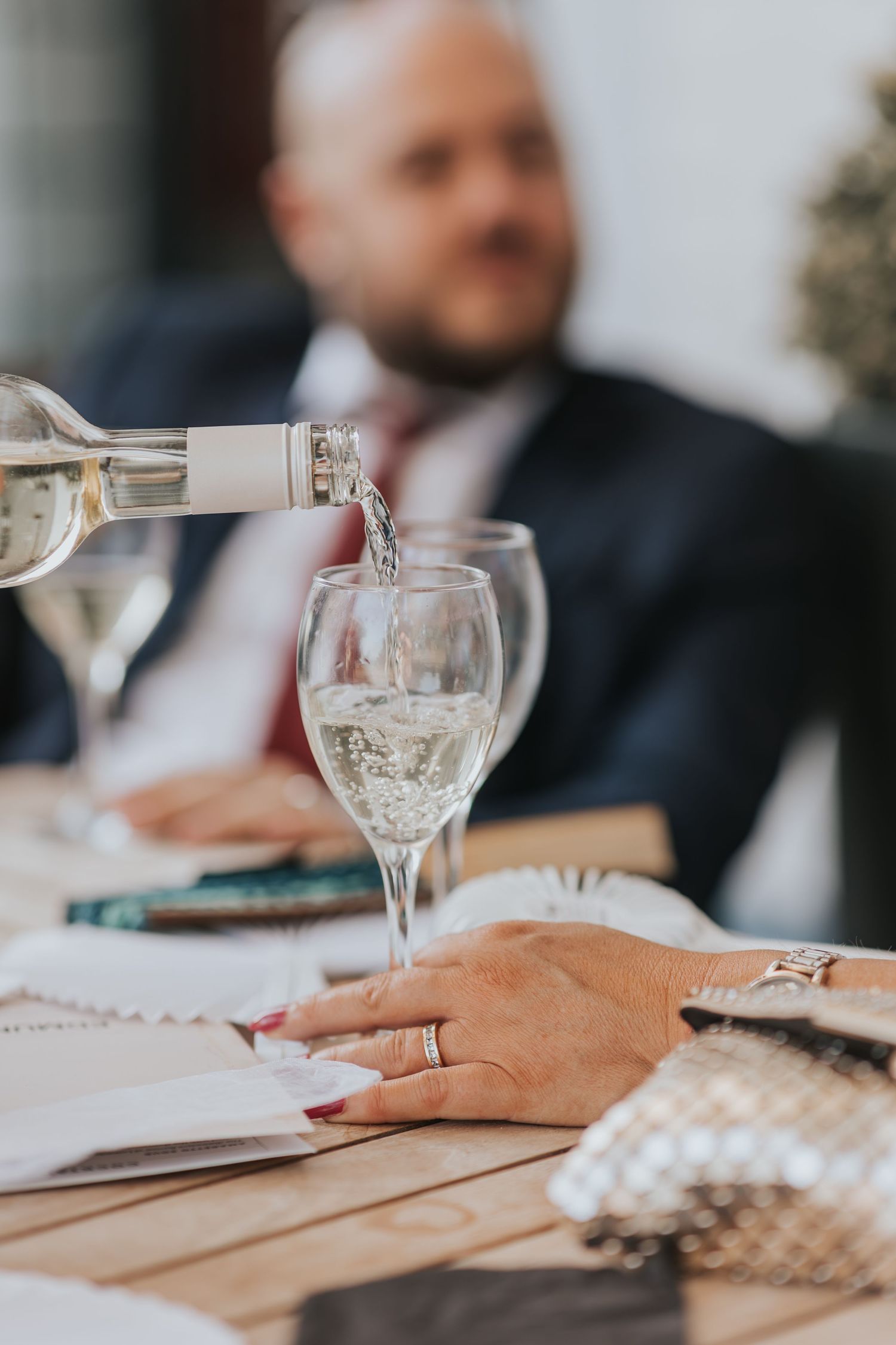 White wine being poured into a glass at a formal dinner table, with a man in a suit sitting in the background.