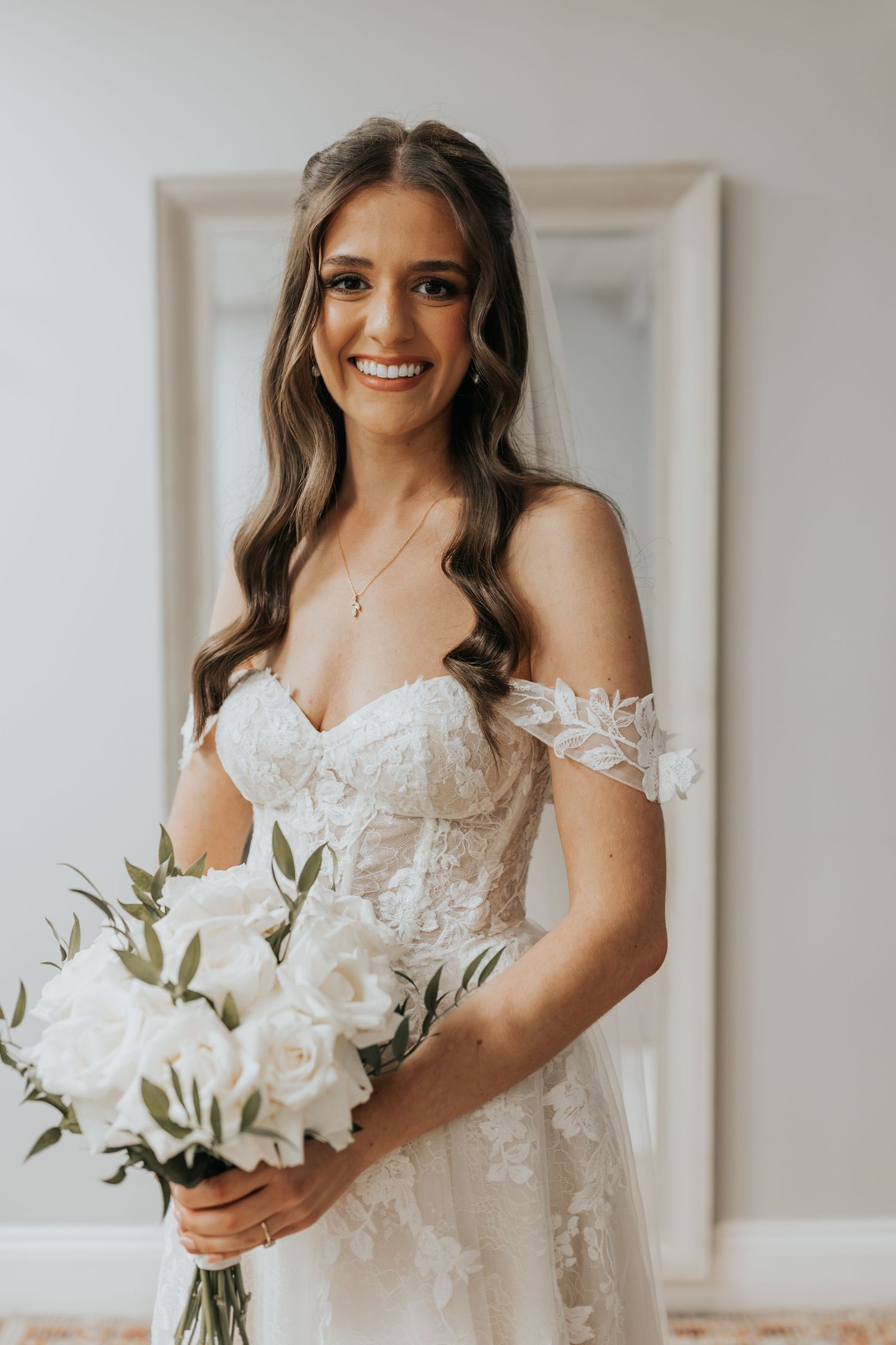 Smiling bride in off-shoulder lace wedding gown holding white rose bouquet with veil, posing indoors.