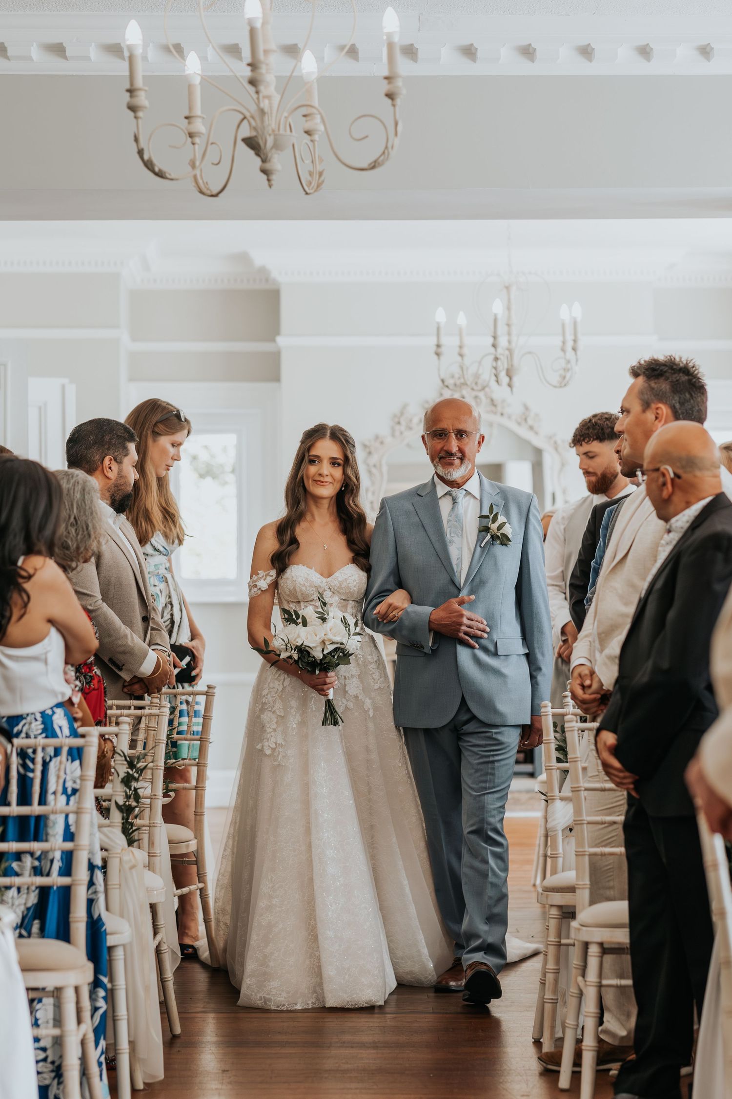 Bride in lace gown walks down aisle with older gentleman in blue suit at elegant indoor wedding ceremony.
