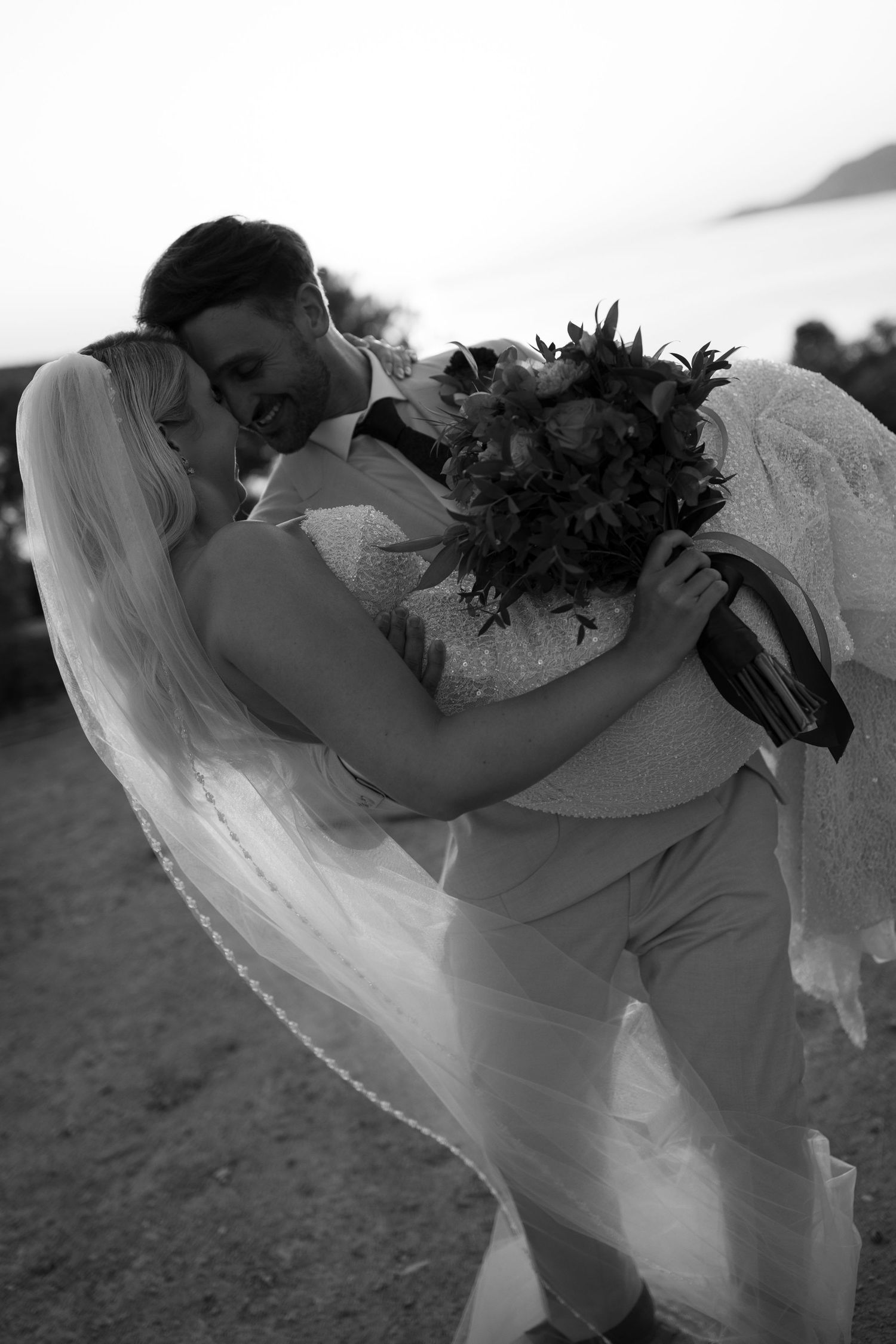 A black and white image of a bride and groom embracing and holding a bouquet of flowers on a field.