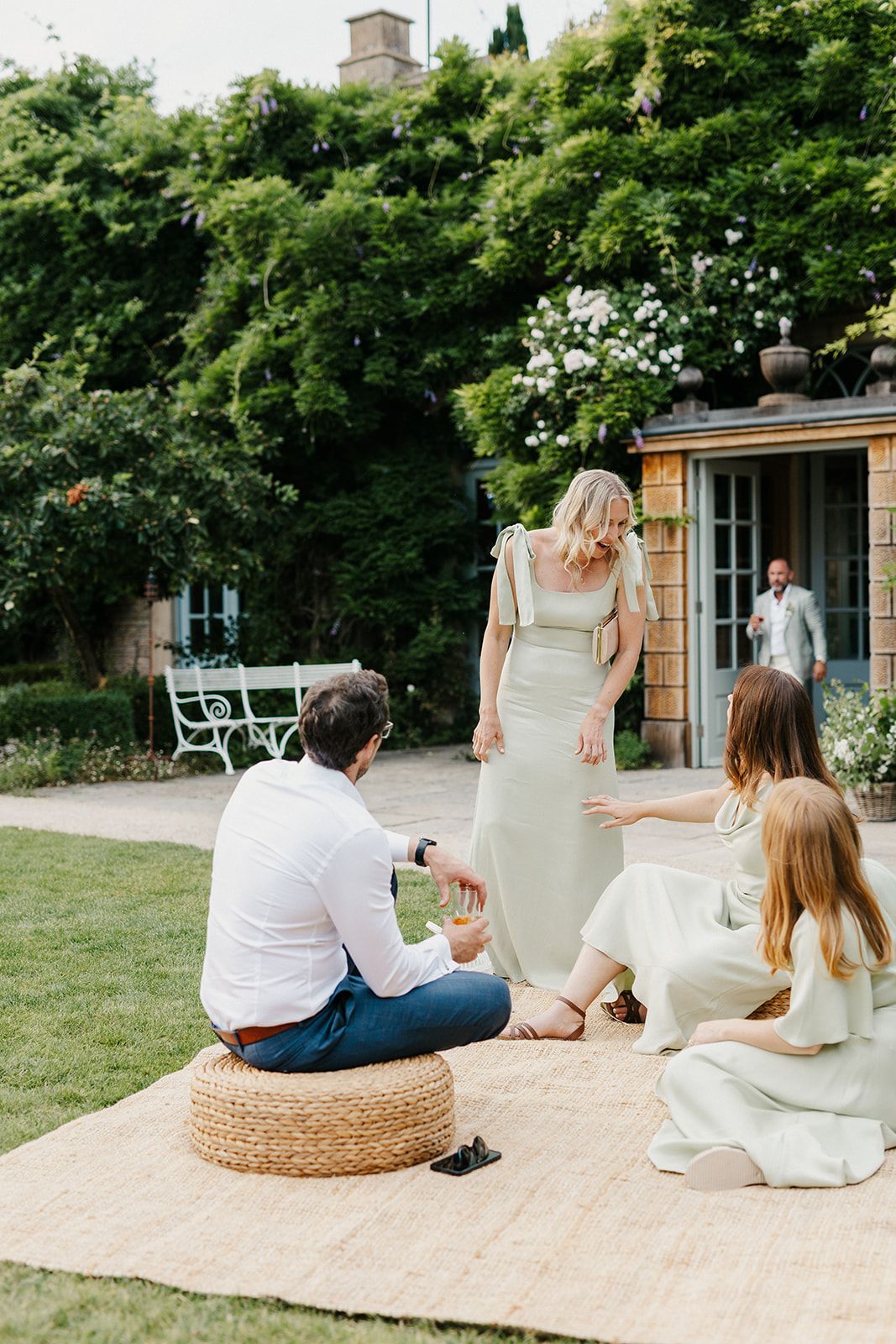Guests relaxing on a woven rug in a lush garden, with a woman in a sage green dress standing nearby.