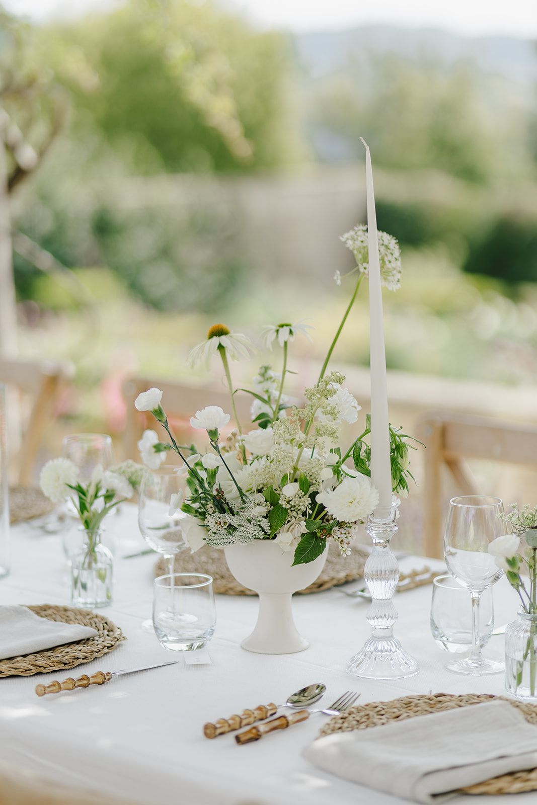 Elegant wedding table setting with white floral centerpiece, taper candle, woven placemats, and glassware in an outdoor venue.