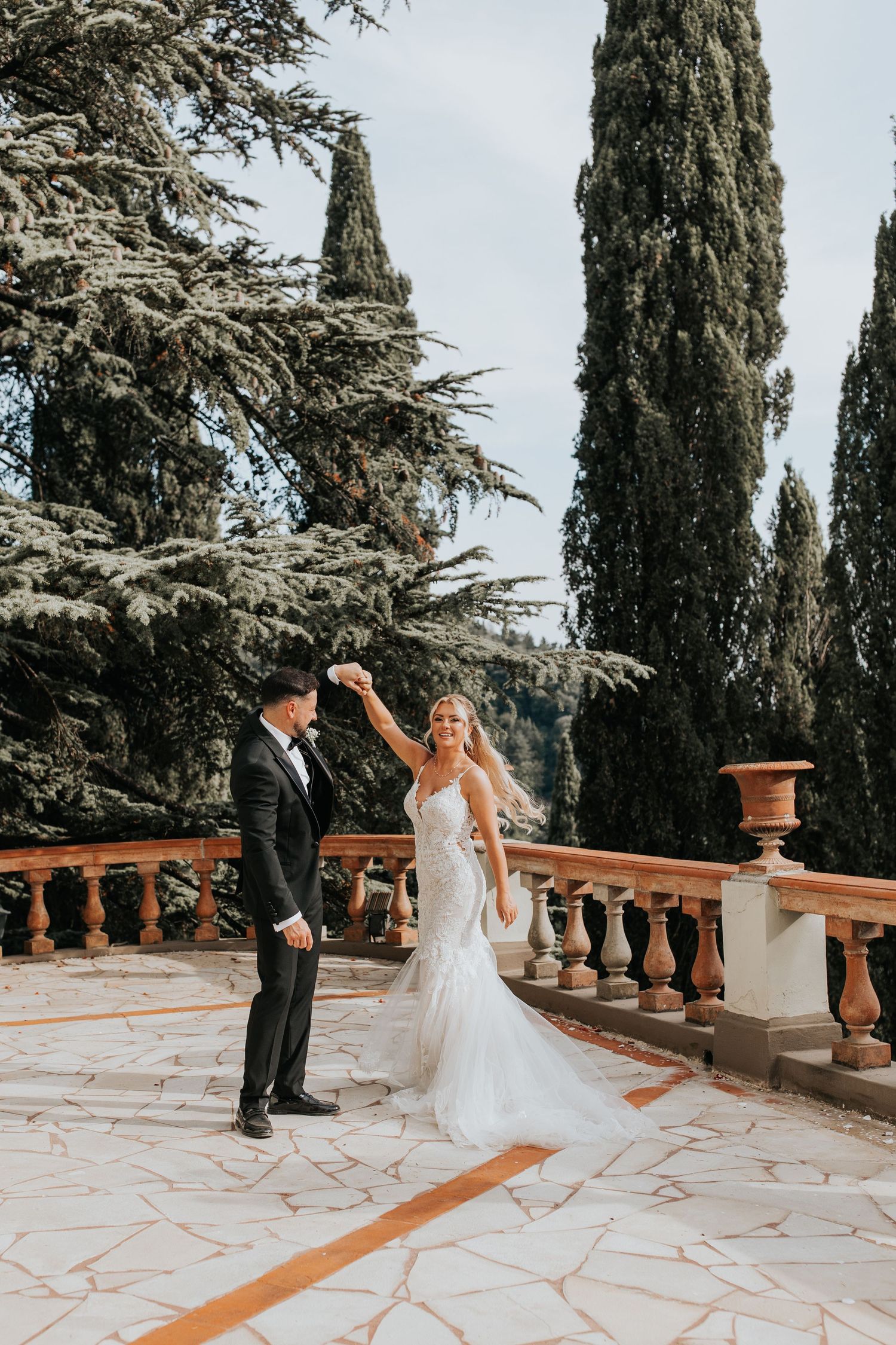 A bride and groom stand on a stone patio surrounded by lush greenery and trees.
