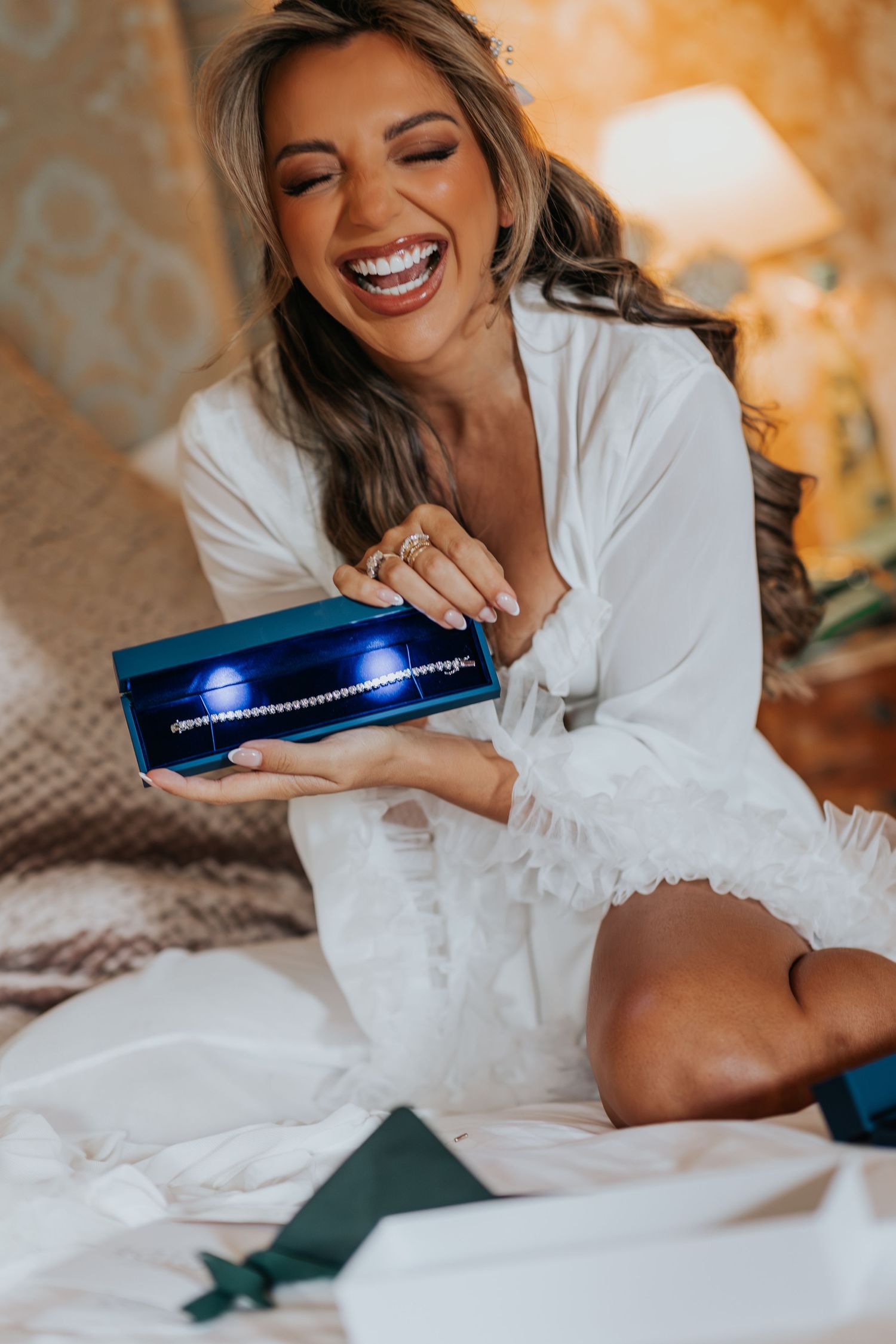 Woman in white robe laughing while opening a blue jewelry box on a bed.