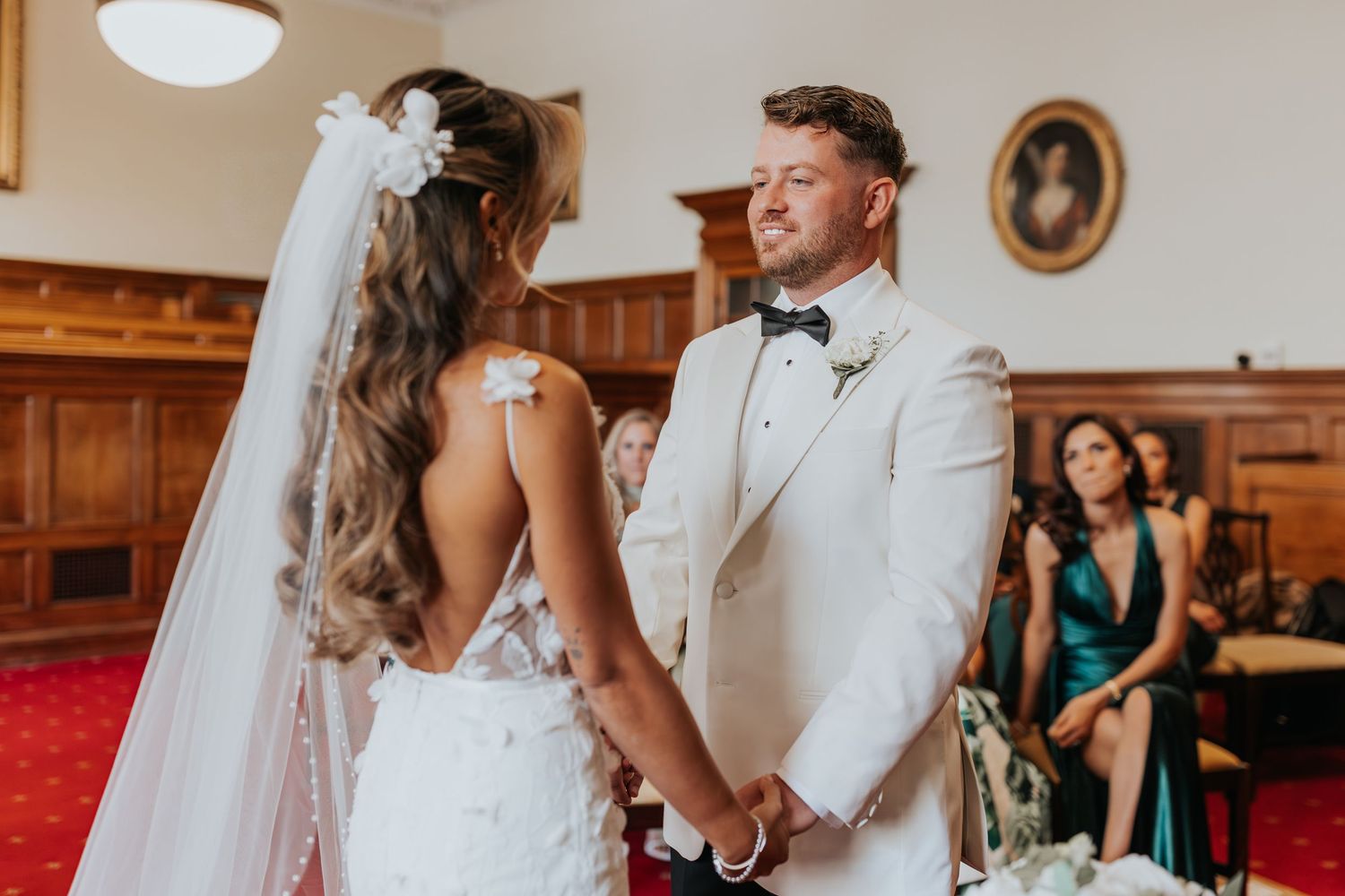 Bride and groom holding hands at wedding ceremony in elegant room with guests seated behind them.