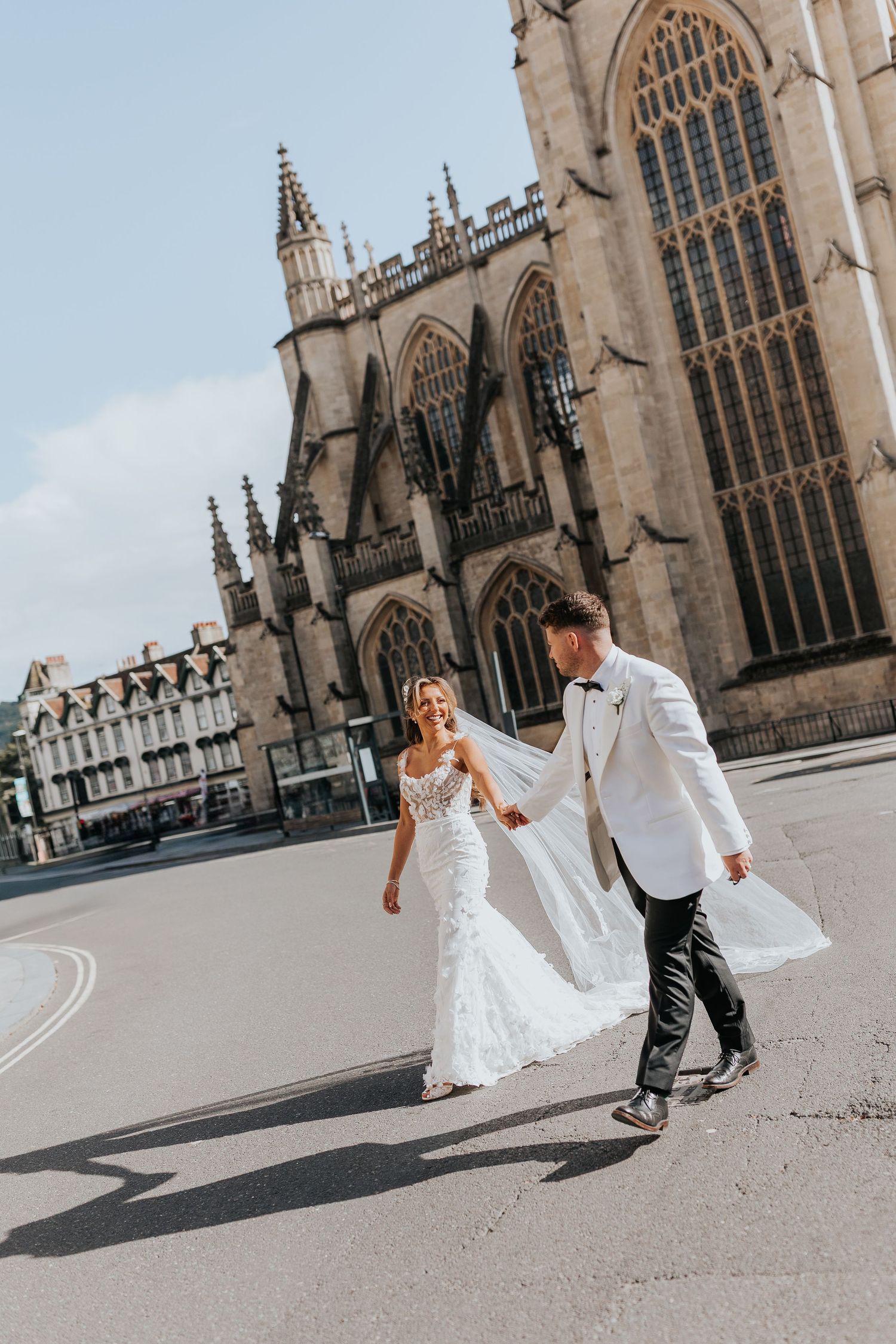 Bride and groom walking hand in hand outside Bath Abbey on their wedding day in elegant white attire.