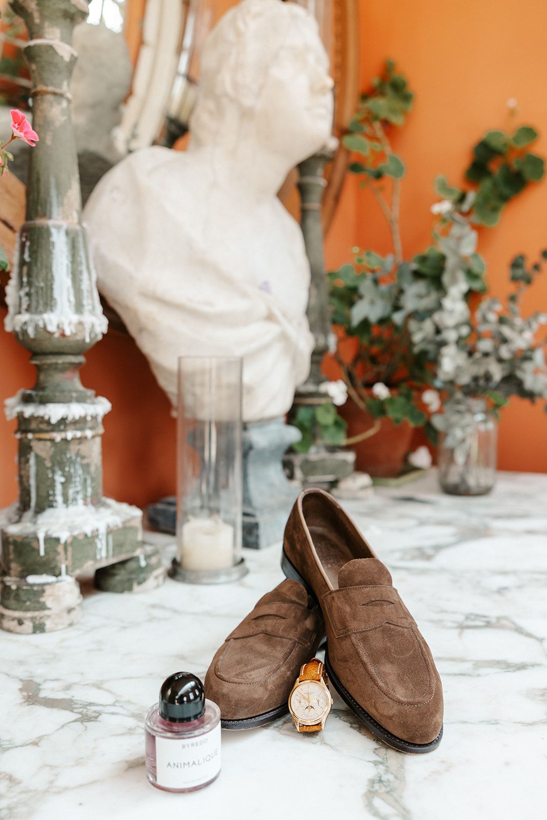 Brown suede loafers with a gold watch and cologne on a marble surface, with a classical bust sculpture in the background.