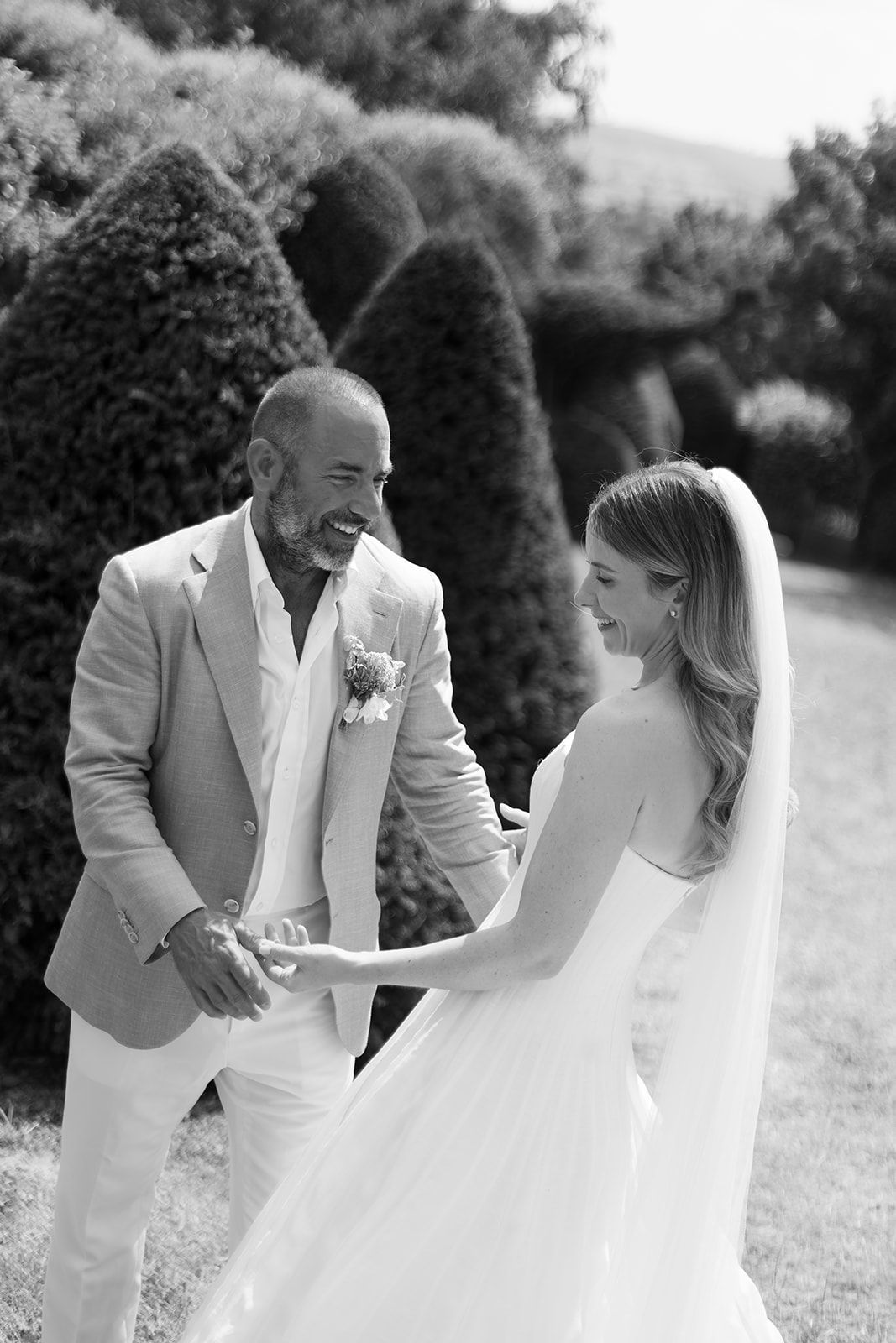 Black and white photo of a smiling groom in a tan suit holding hands with his bride in a white gown in a garden.