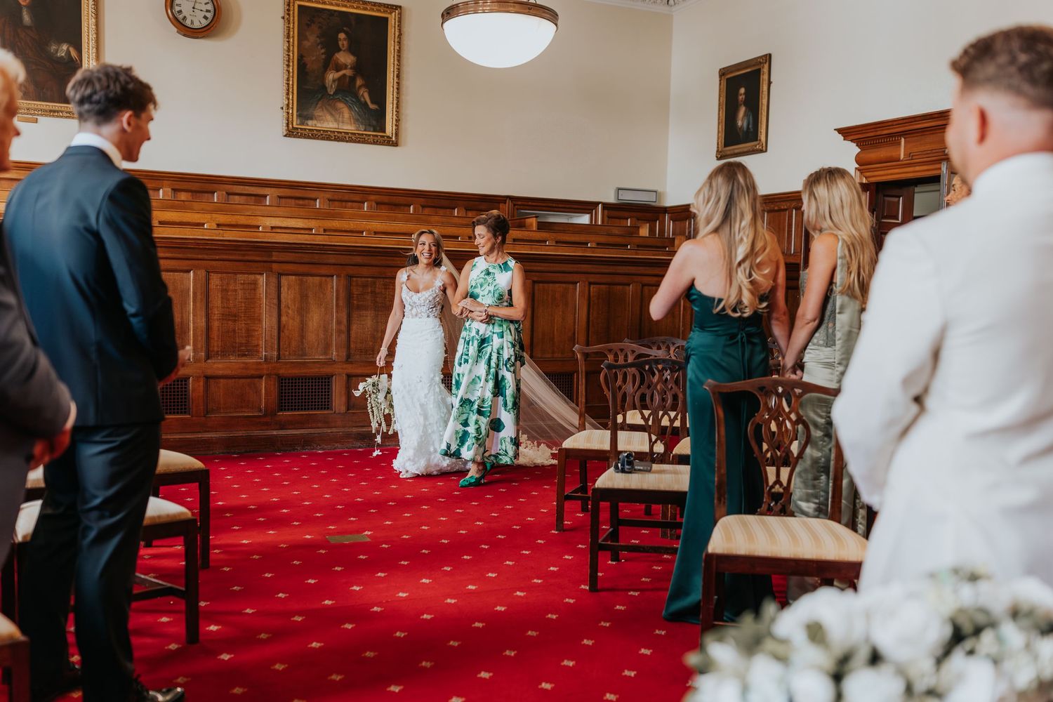 Bride walking down the aisle in a floral gown at an elegant wedding ceremony in a wood-paneled hall.