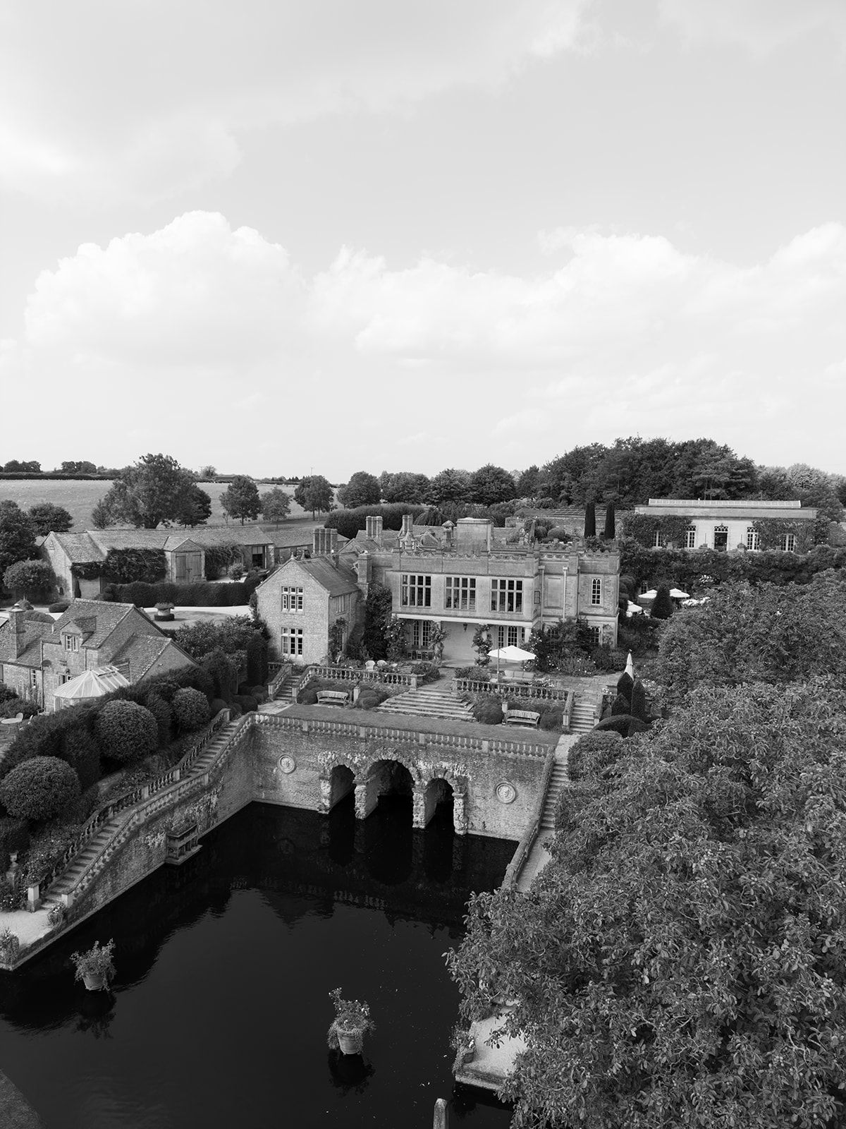 Aerial black and white view of a historic English manor house with arched bridge over a moat surrounded by lush trees.