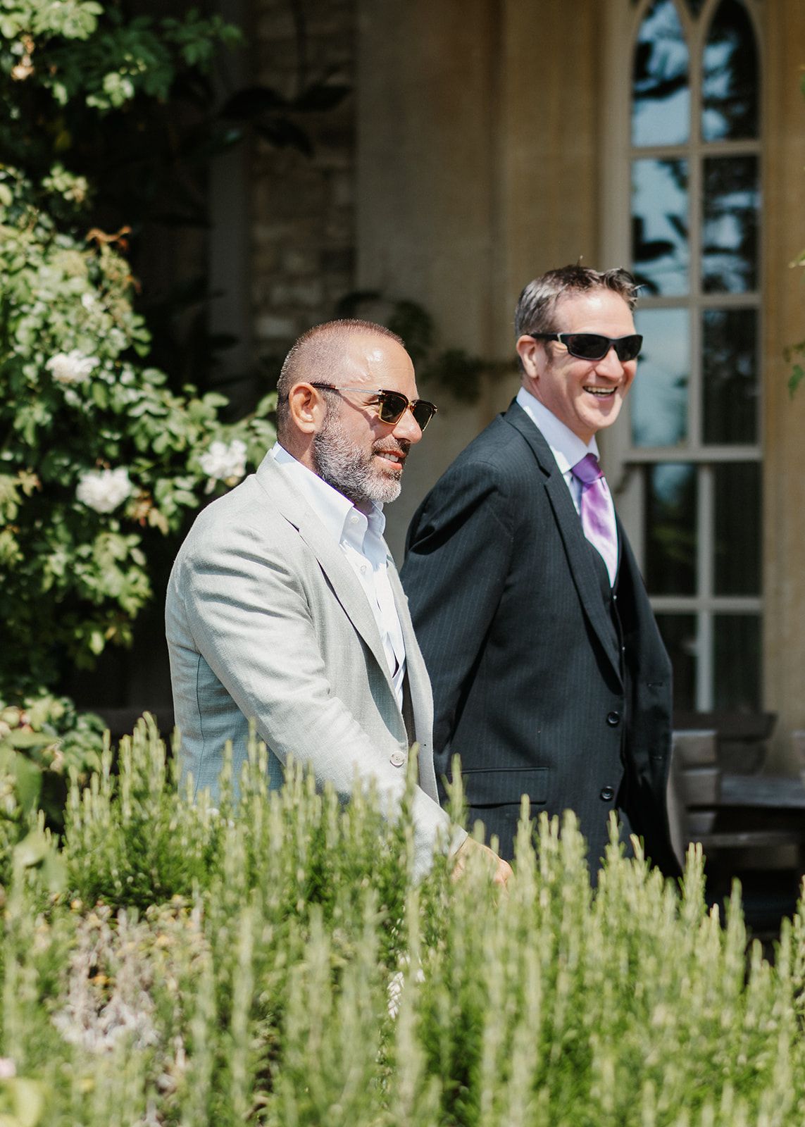 Two men in suits and sunglasses smiling outdoors at a garden wedding venue on a sunny day.