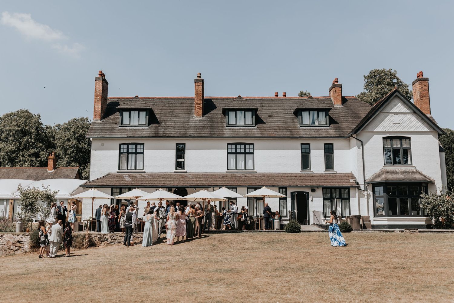 Guests gather outside a white country house venue on a sunny day for an outdoor wedding reception.