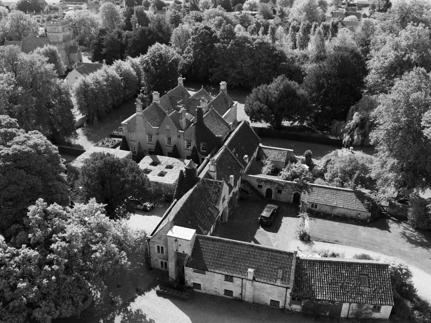 Aerial black and white photo of a historic manor house surrounded by dense trees in a rural European countryside setting.