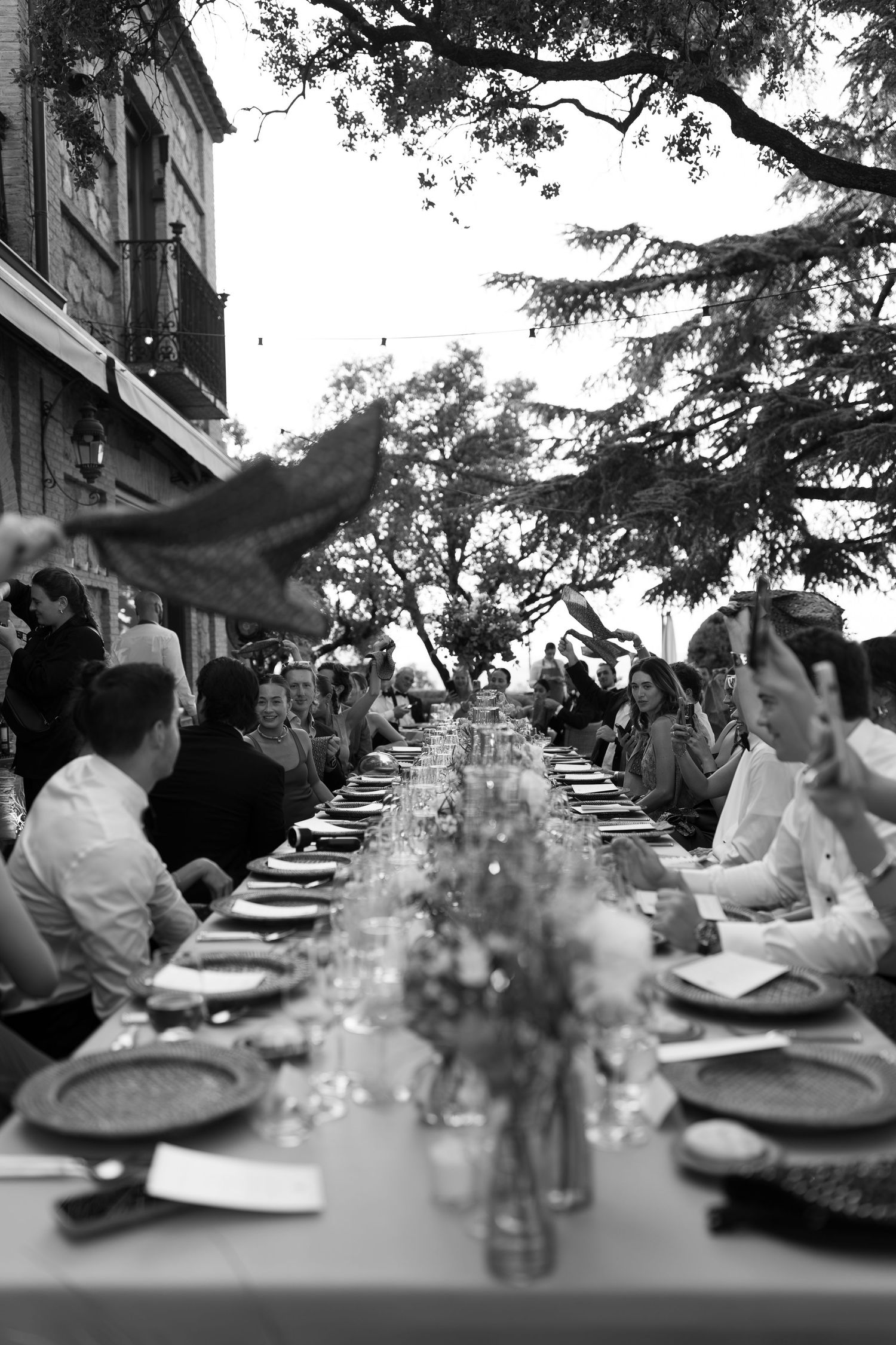 A black and white image of a long table set for a formal outdoor dinner party under the shade of trees.