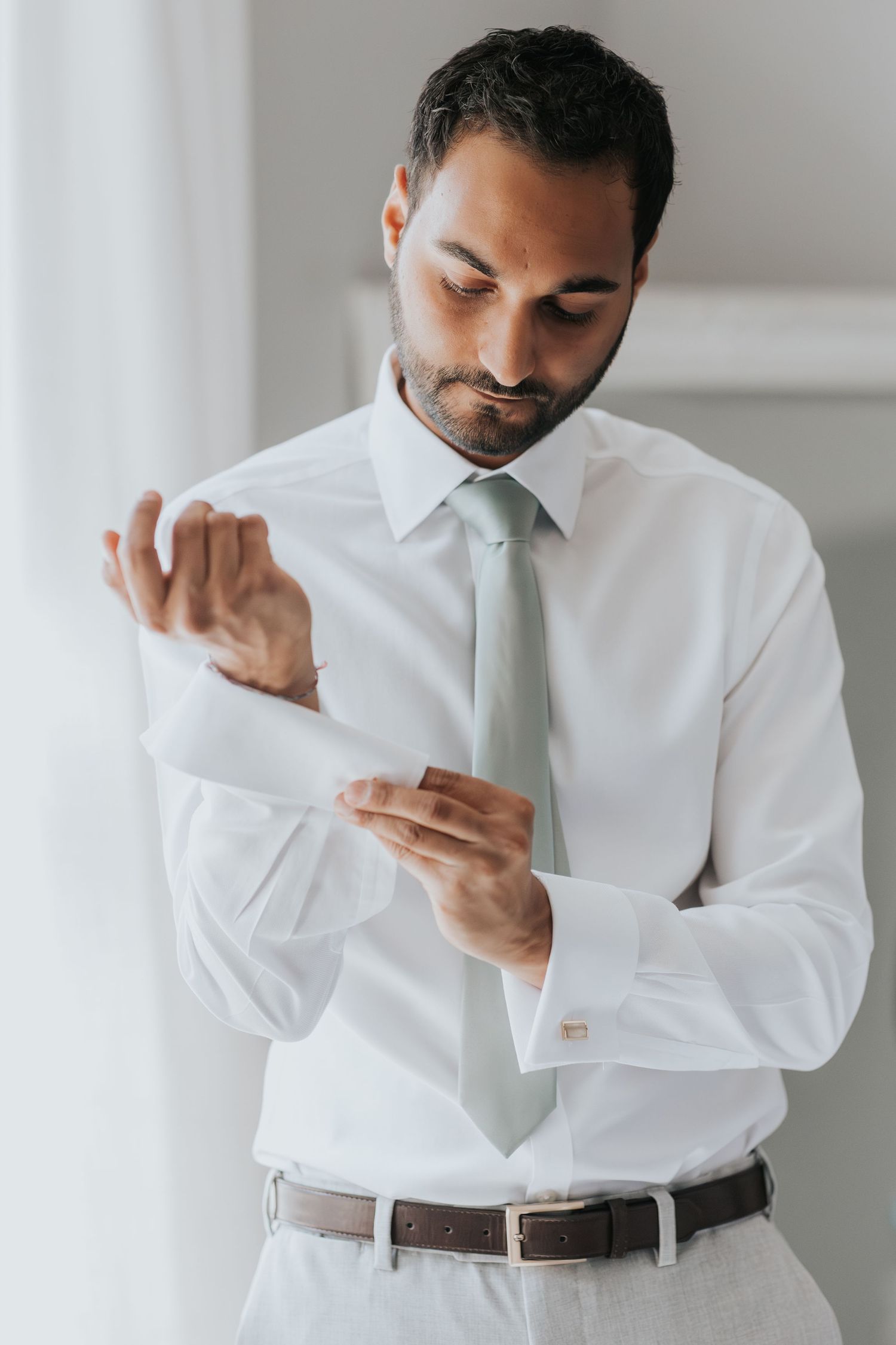 Man in white dress shirt and light tie fastening cufflinks, dressed for a formal occasion.