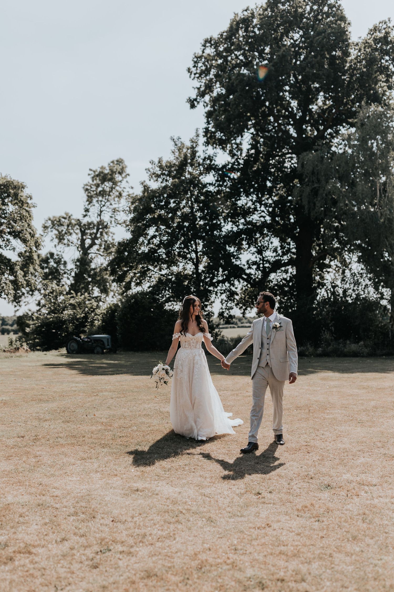 Bride and groom walk hand in hand across a sunny green field with large trees in the background.