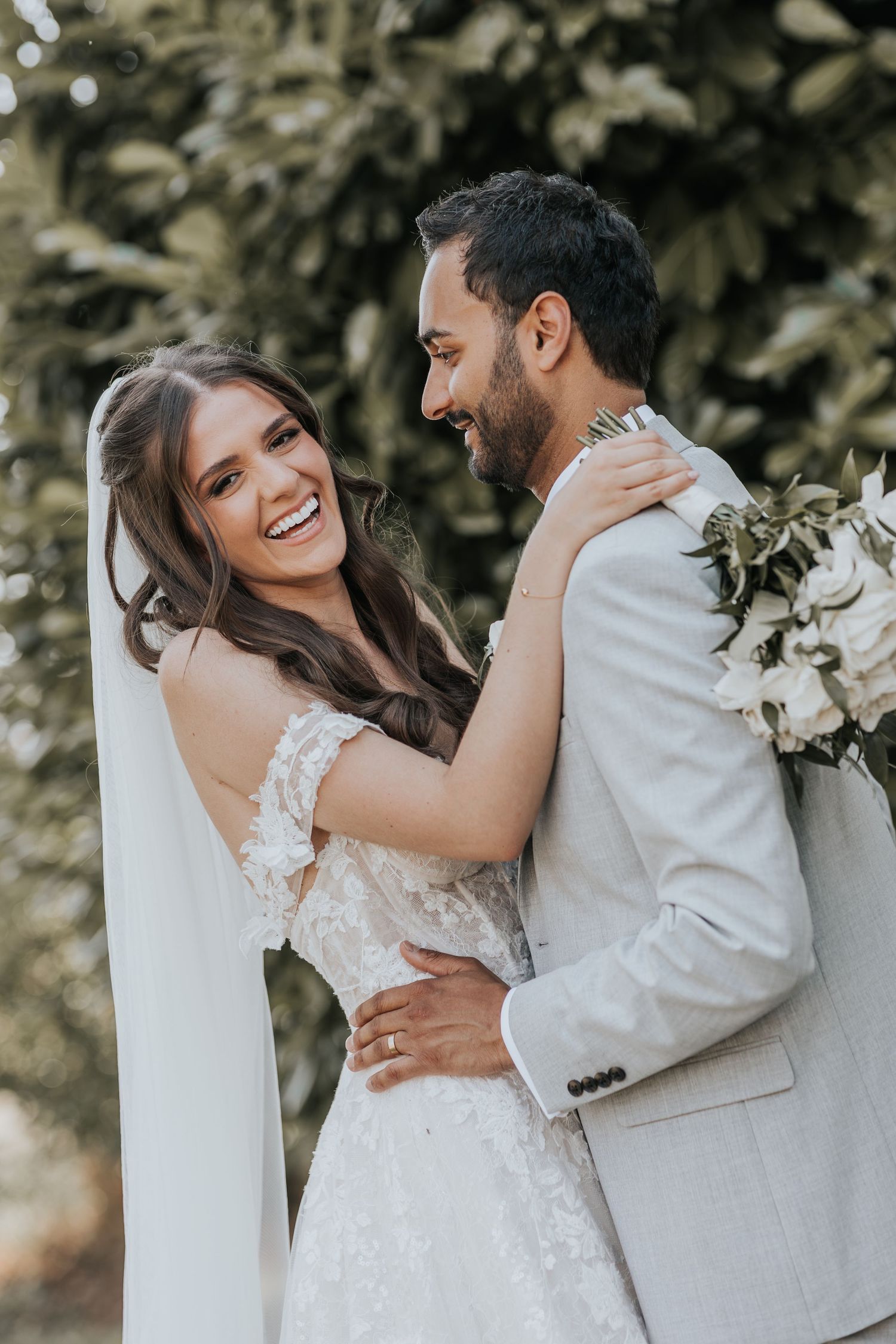 A joyful bride and groom embrace outdoors, smiling at each other, she in a lace gown with veil, he in a light gray suit.