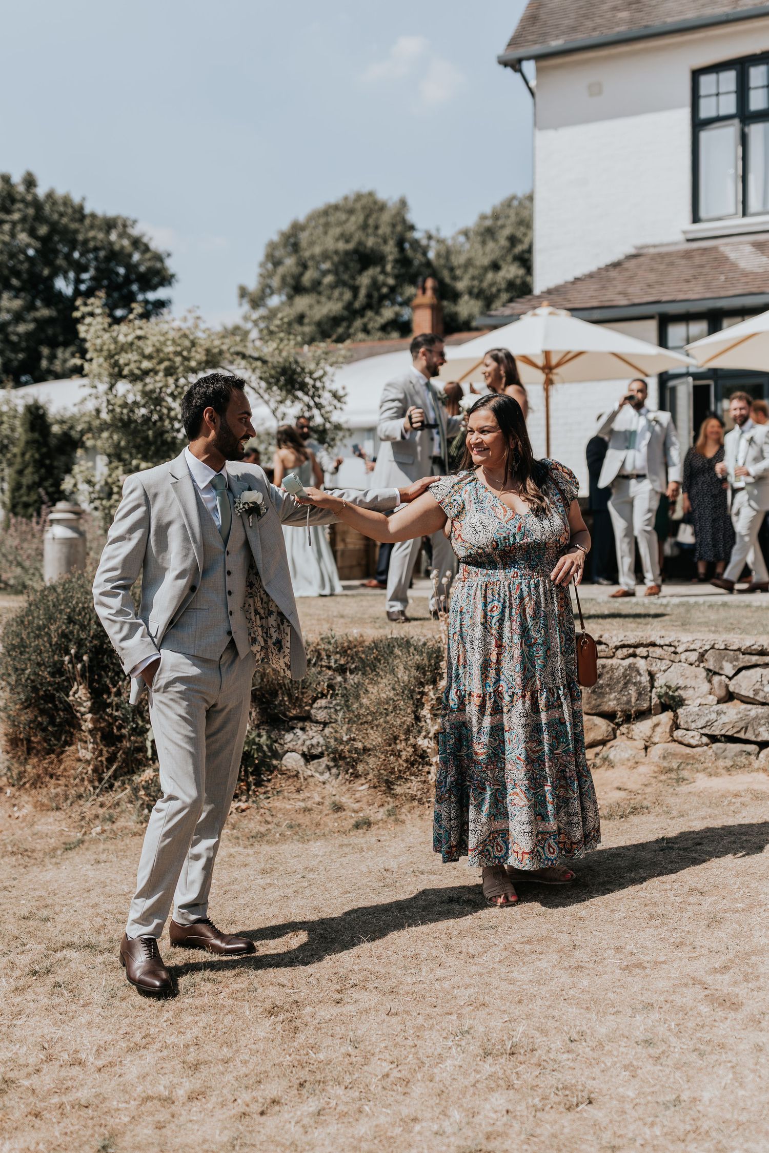 A couple dances outdoors at a sunny wedding venue, man in grey suit, woman in floral maxi dress.