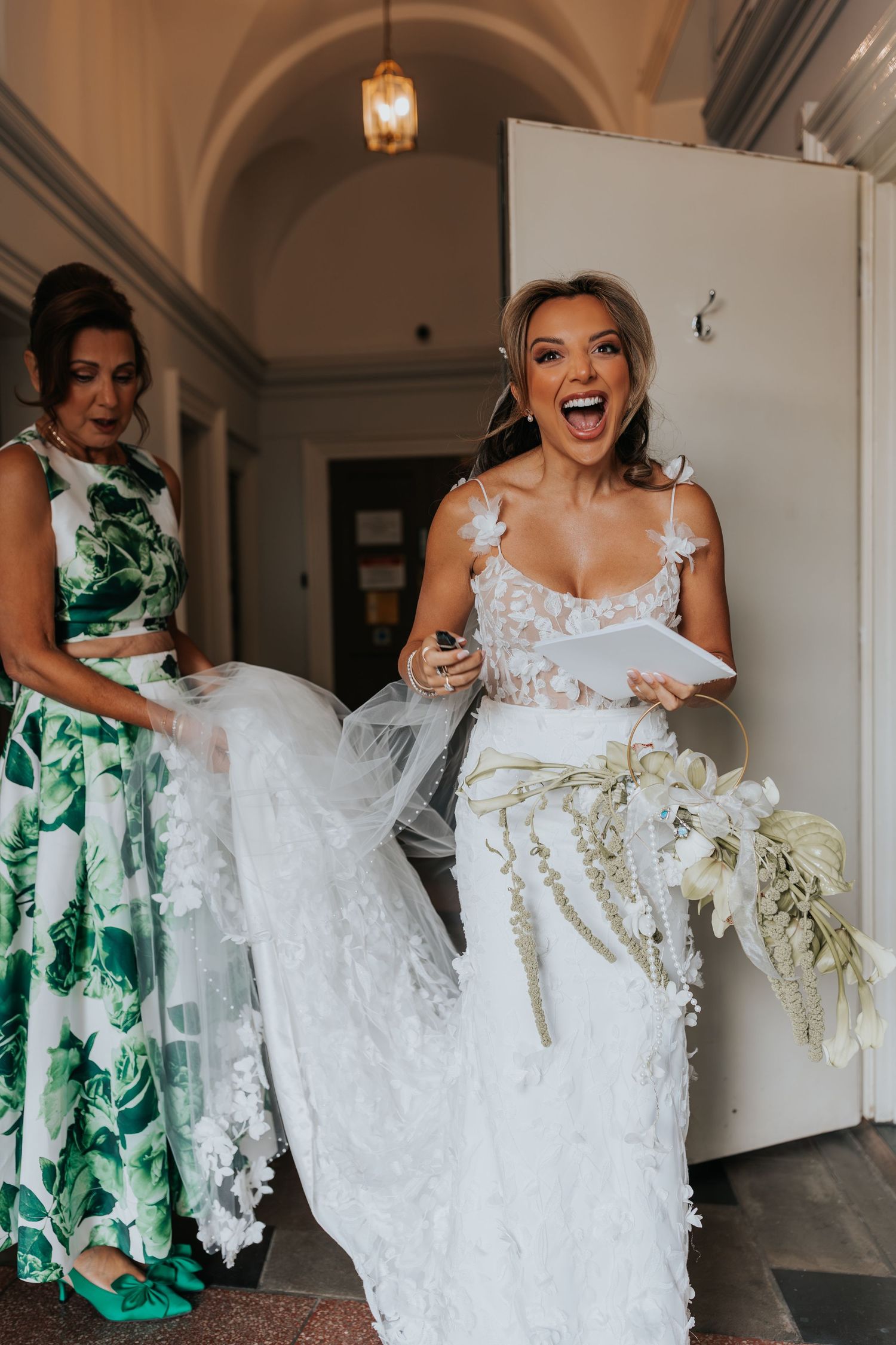 Excited bride in lace wedding dress holds bouquet while bridesmaid in green floral dress assists with veil in hallway.