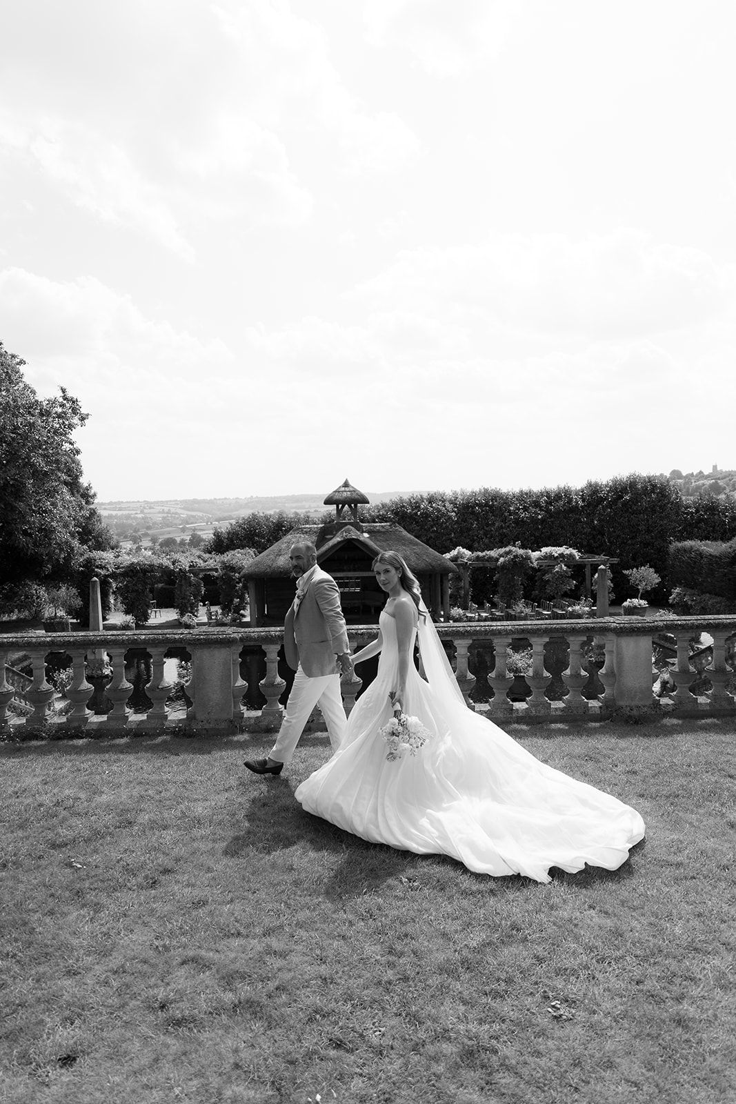 Black and white photo of a bride and groom walking hand in hand through a formal garden with stone balustrade.