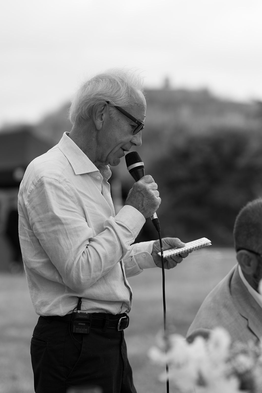 Elderly man with glasses reading from notepad while speaking into microphone outdoors, black and white photo.