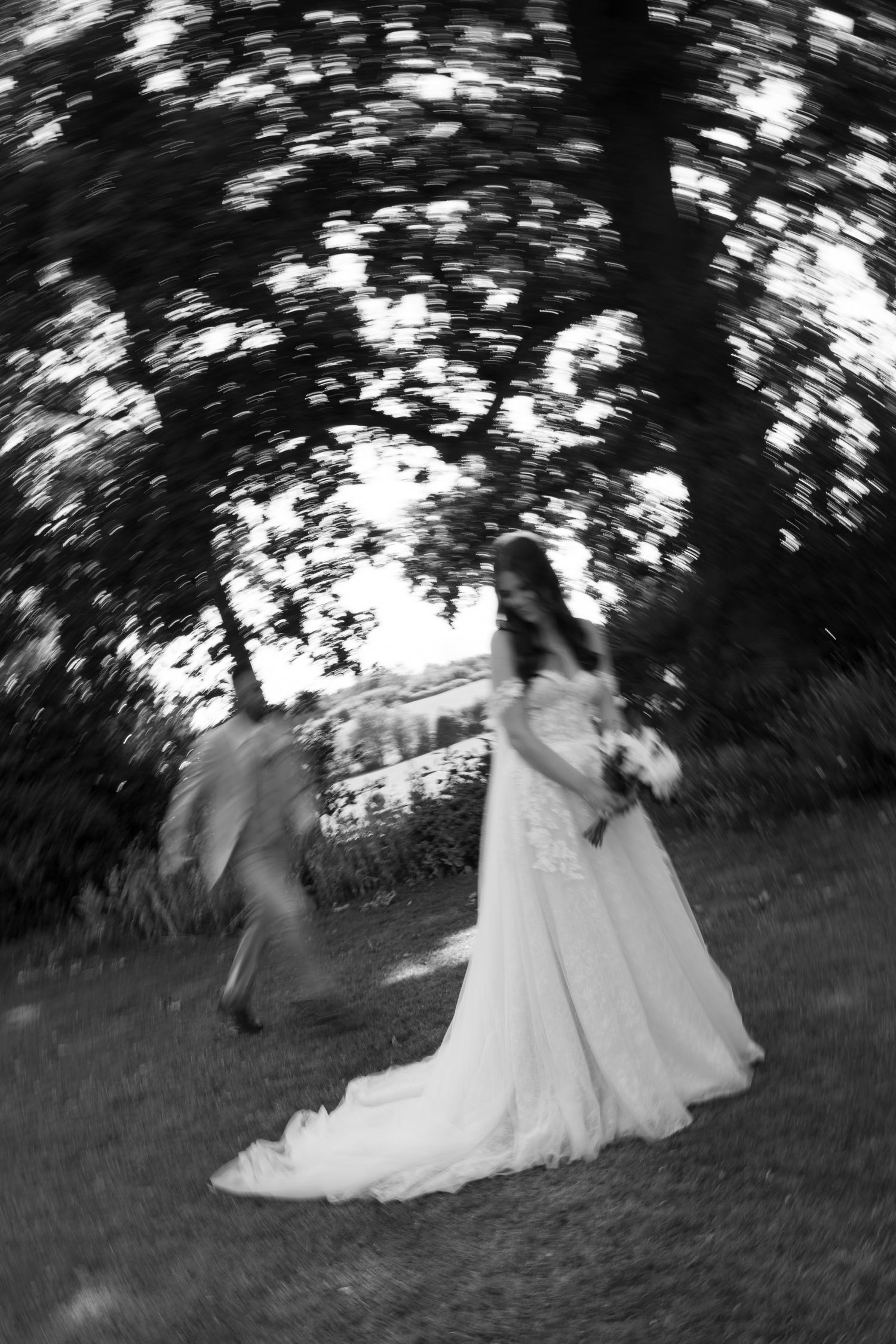 Black and white photo of a bride in a white gown holding bouquet outdoors, with motion blur effect under trees.