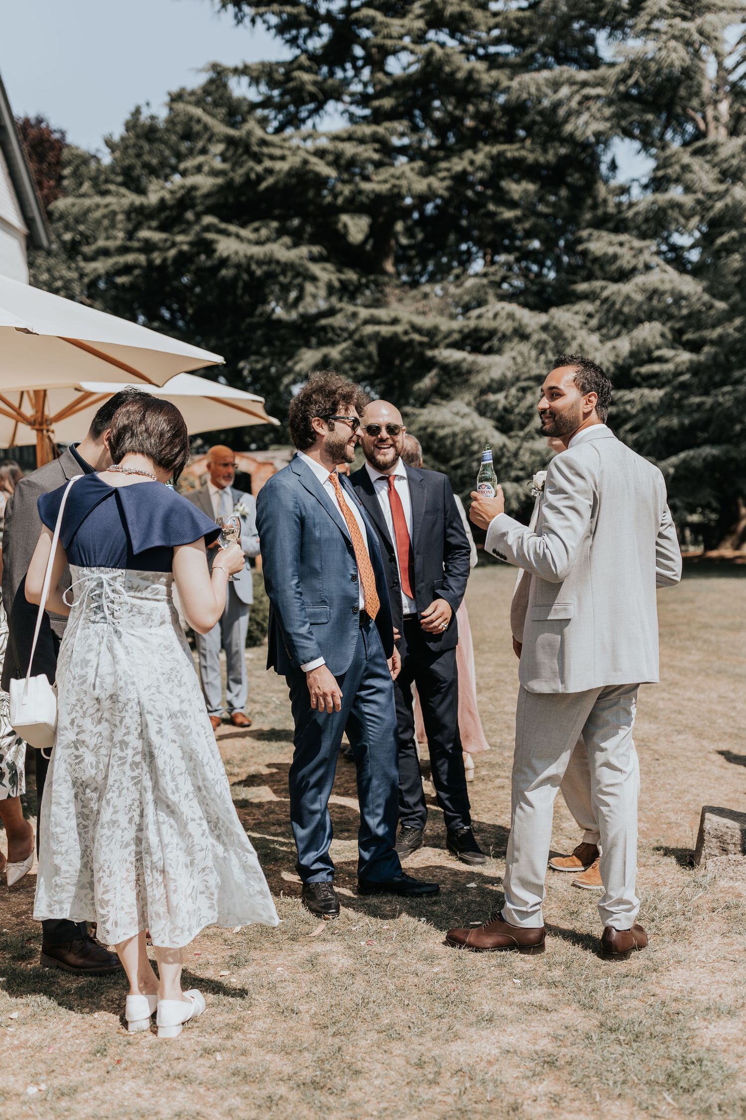Guests socializing outdoors at a summer wedding reception, men in suits holding drinks on a sunny lawn.