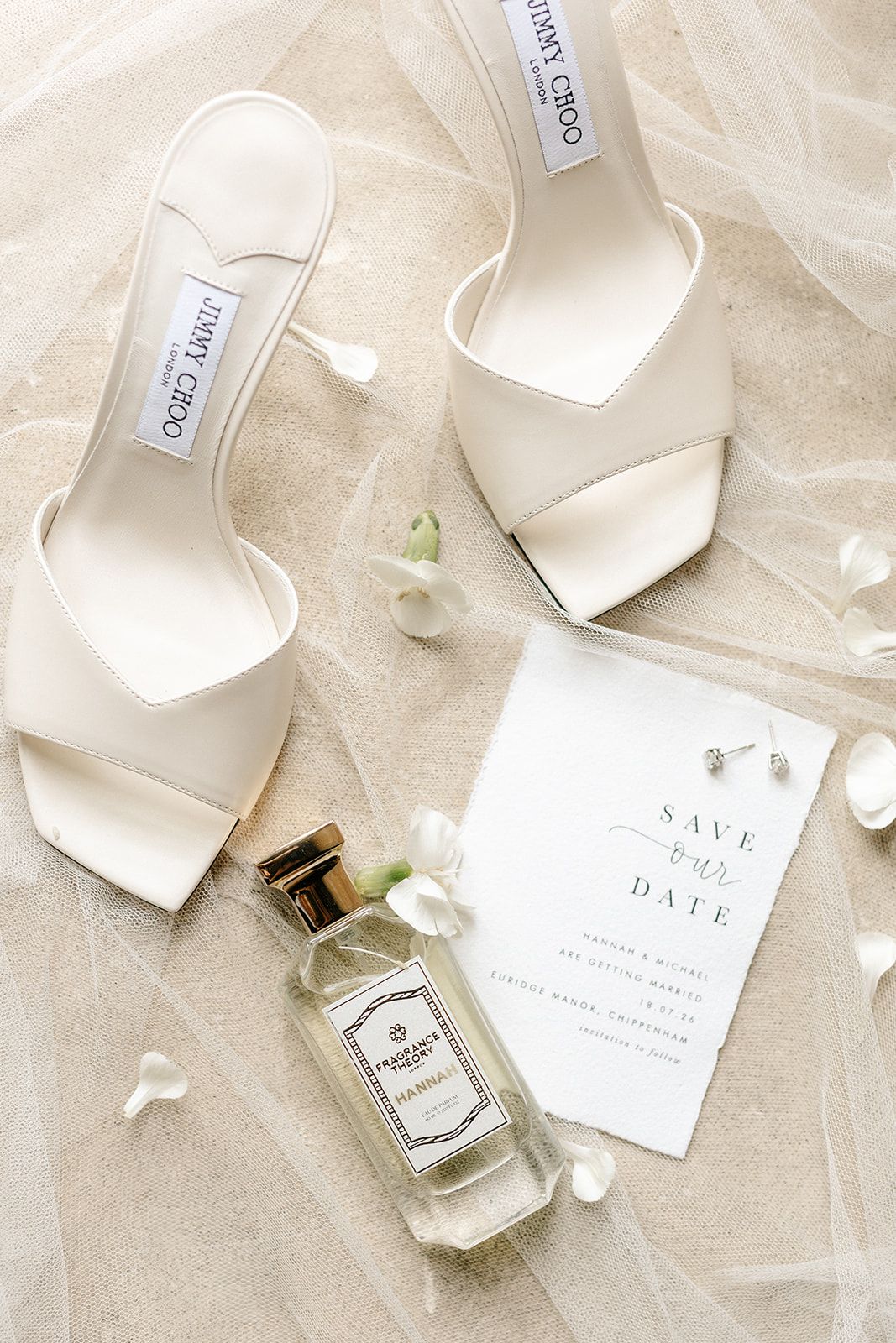 White Jimmy Choo bridal heels beside a Save the Date card, perfume bottle, and flowers on a veil.
