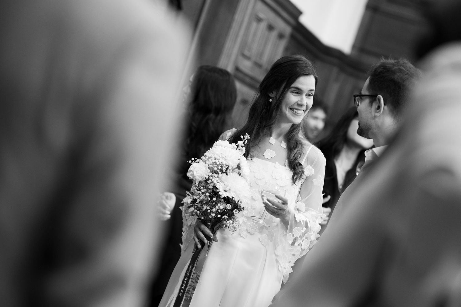 A woman in a white wedding dress stands among a crowd, holding a bouquet of flowers.