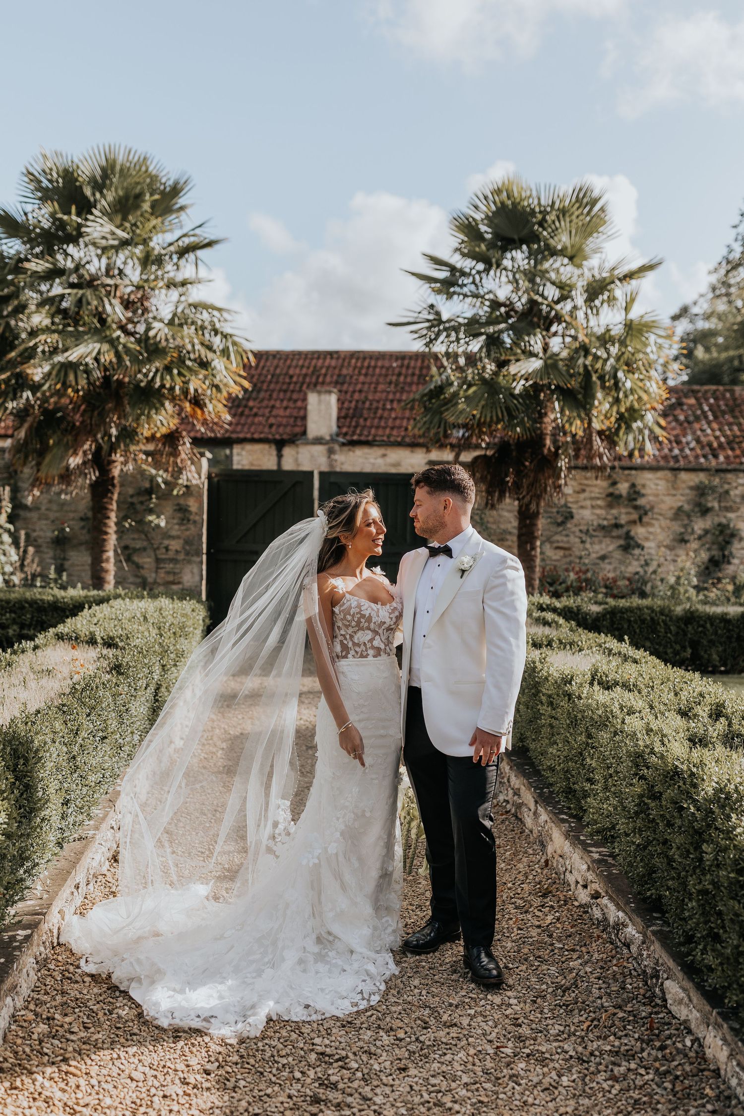 Bride and groom share a romantic moment in a formal garden with palm trees and stone building backdrop on their wedding day.