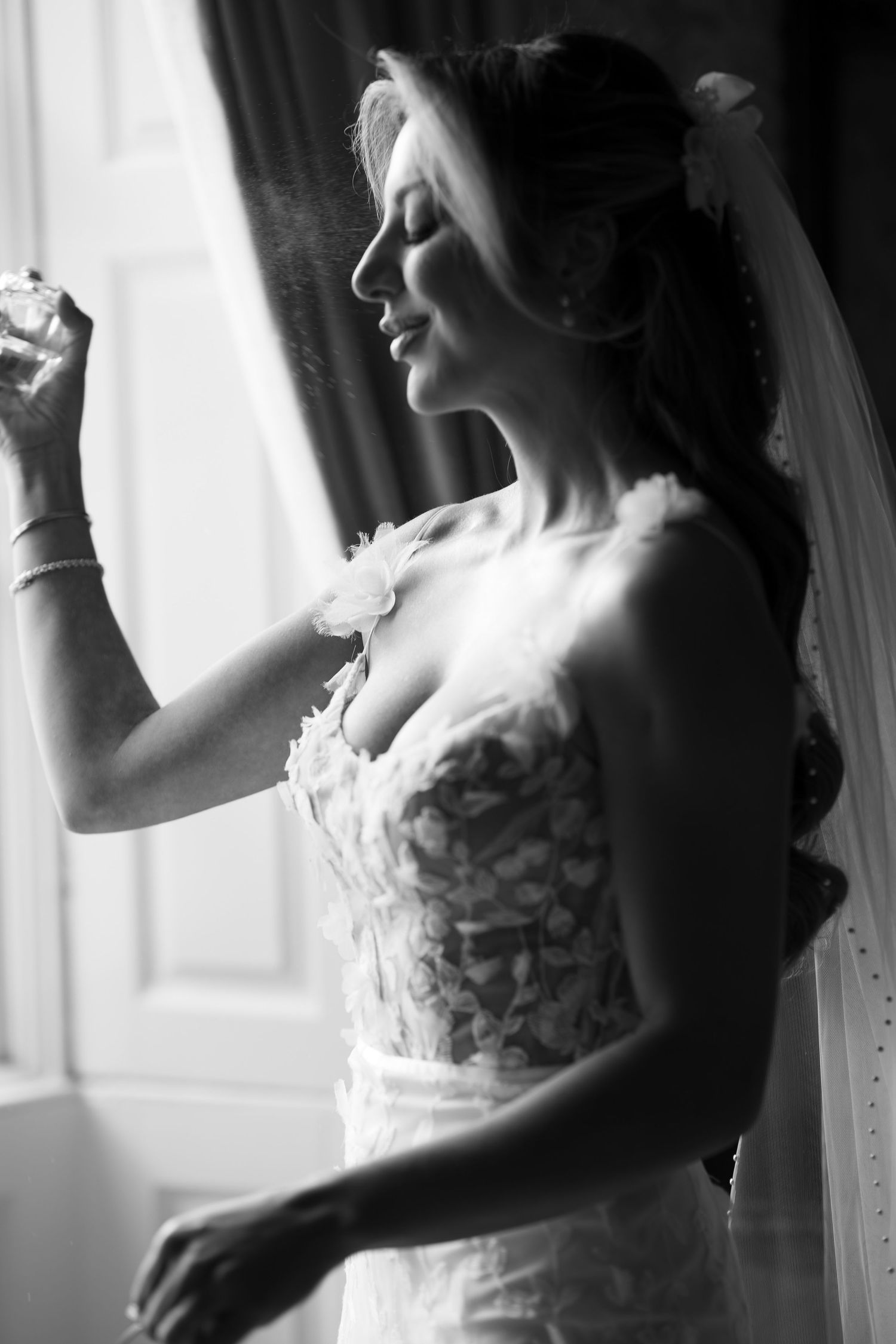 Black and white photo of a bride in a lace wedding dress spraying perfume by a window.