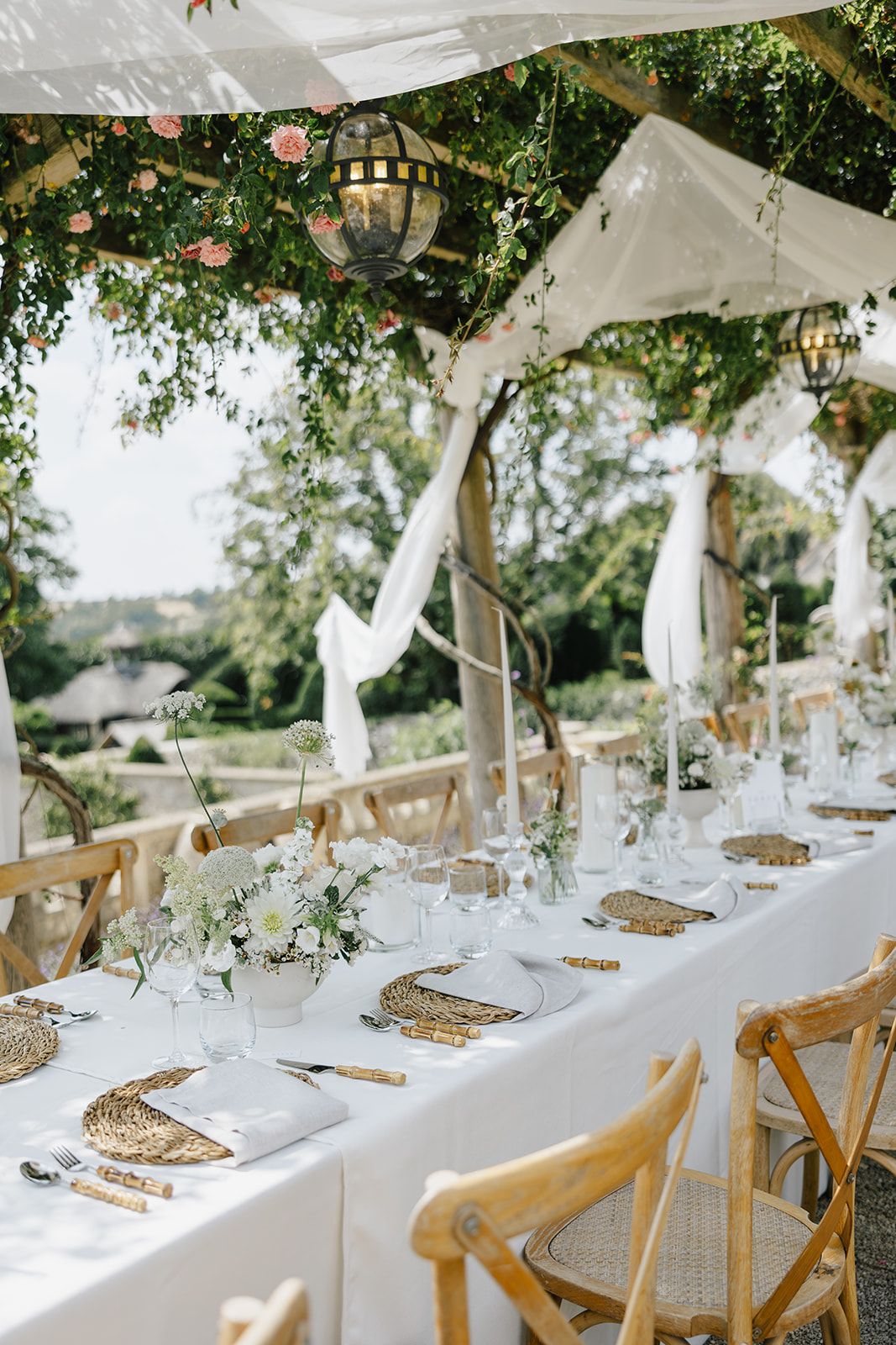 Elegant outdoor wedding reception with long white table, wicker chargers, floral centerpieces, and lanterns under a draped tent.