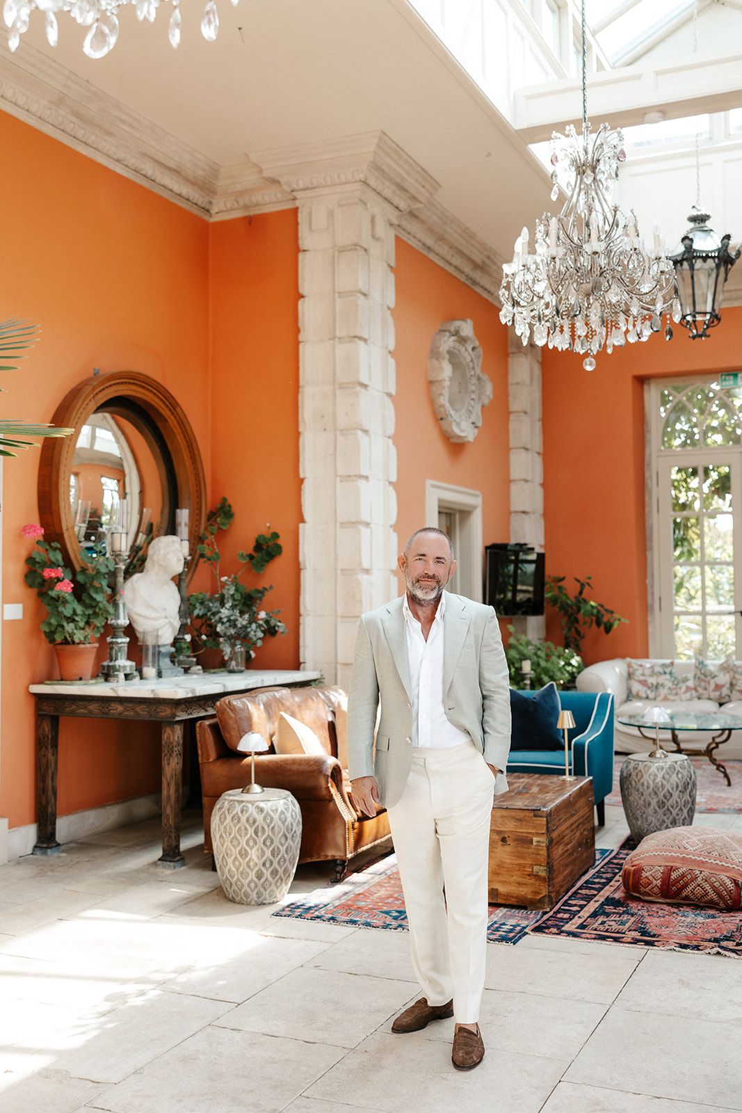 Man in beige suit standing in elegant orange-walled room with chandelier, antique furniture, and colorful rugs.