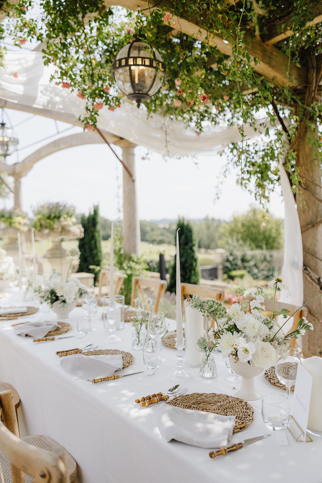 Elegant outdoor wedding reception table with white linens, woven placemats, floral arrangements, and a pergola draped in greenery.