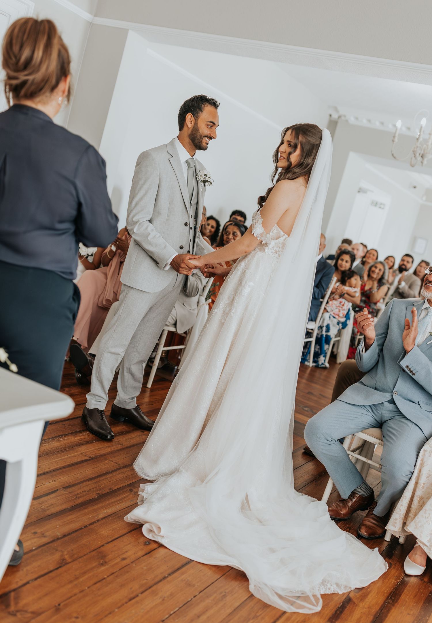 Bride and groom exchange vows at indoor wedding ceremony, guests seated in background, bride in white gown with veil.