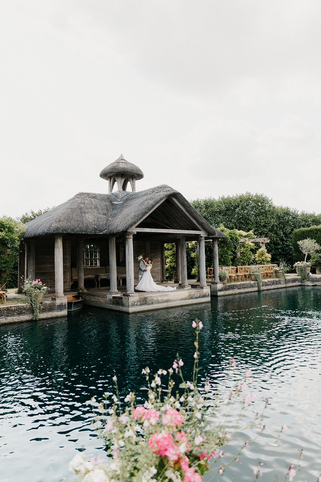 Bride and groom embracing in a thatched gazebo over a serene pond, surrounded by lush gardens and pink flowers.