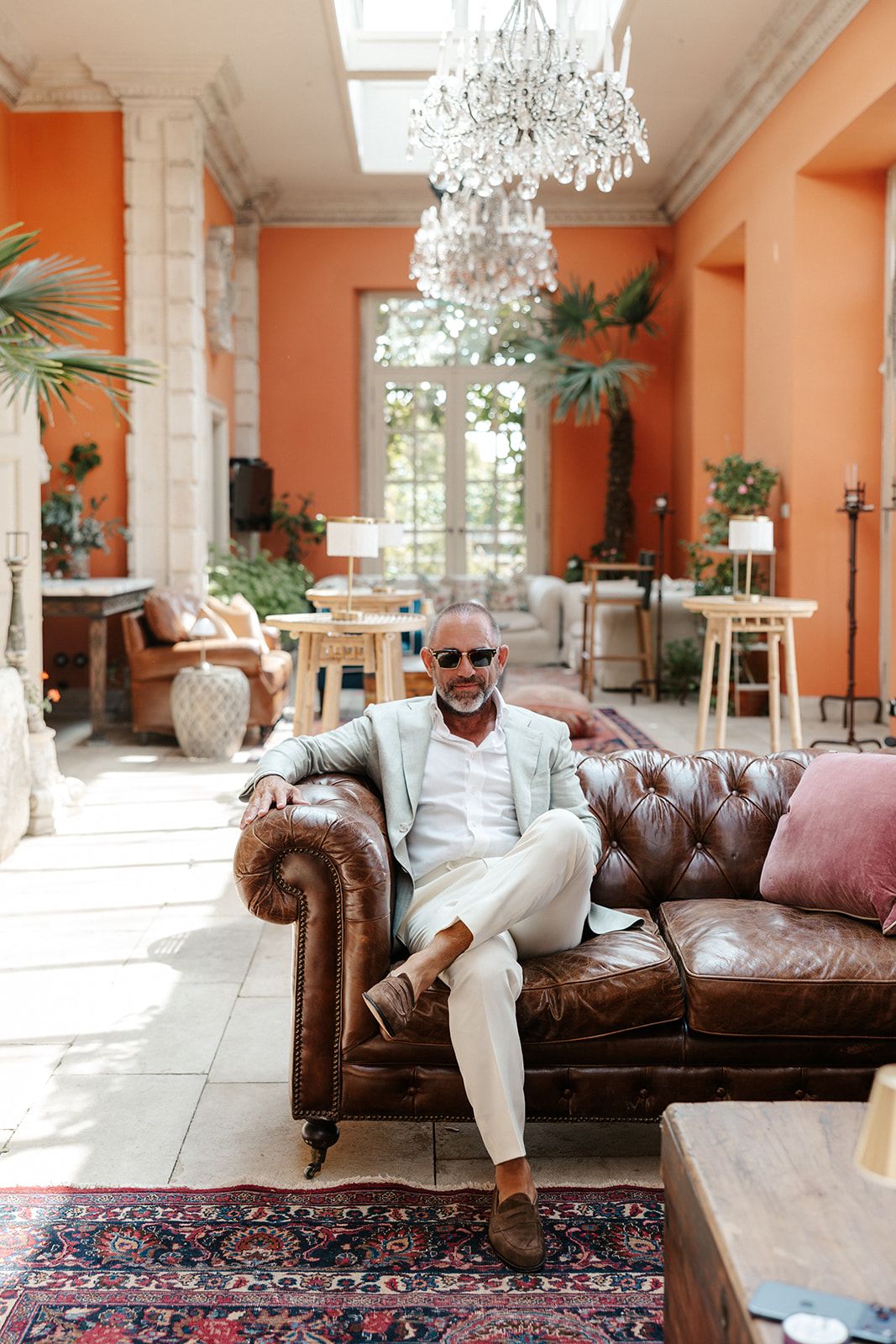 Stylish middle-aged man in white suit and sunglasses relaxing on brown leather Chesterfield sofa in elegant orange room.