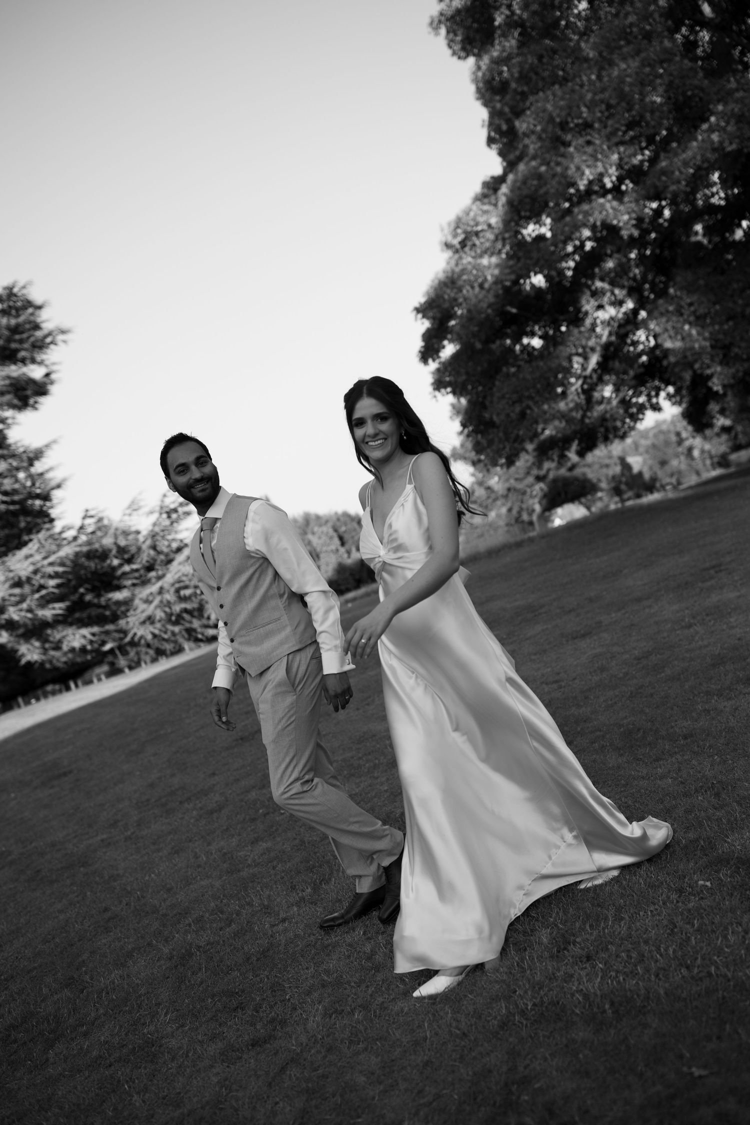 Black and white photo of a bride in a flowing gown and groom in a suit walking on a lush green lawn.