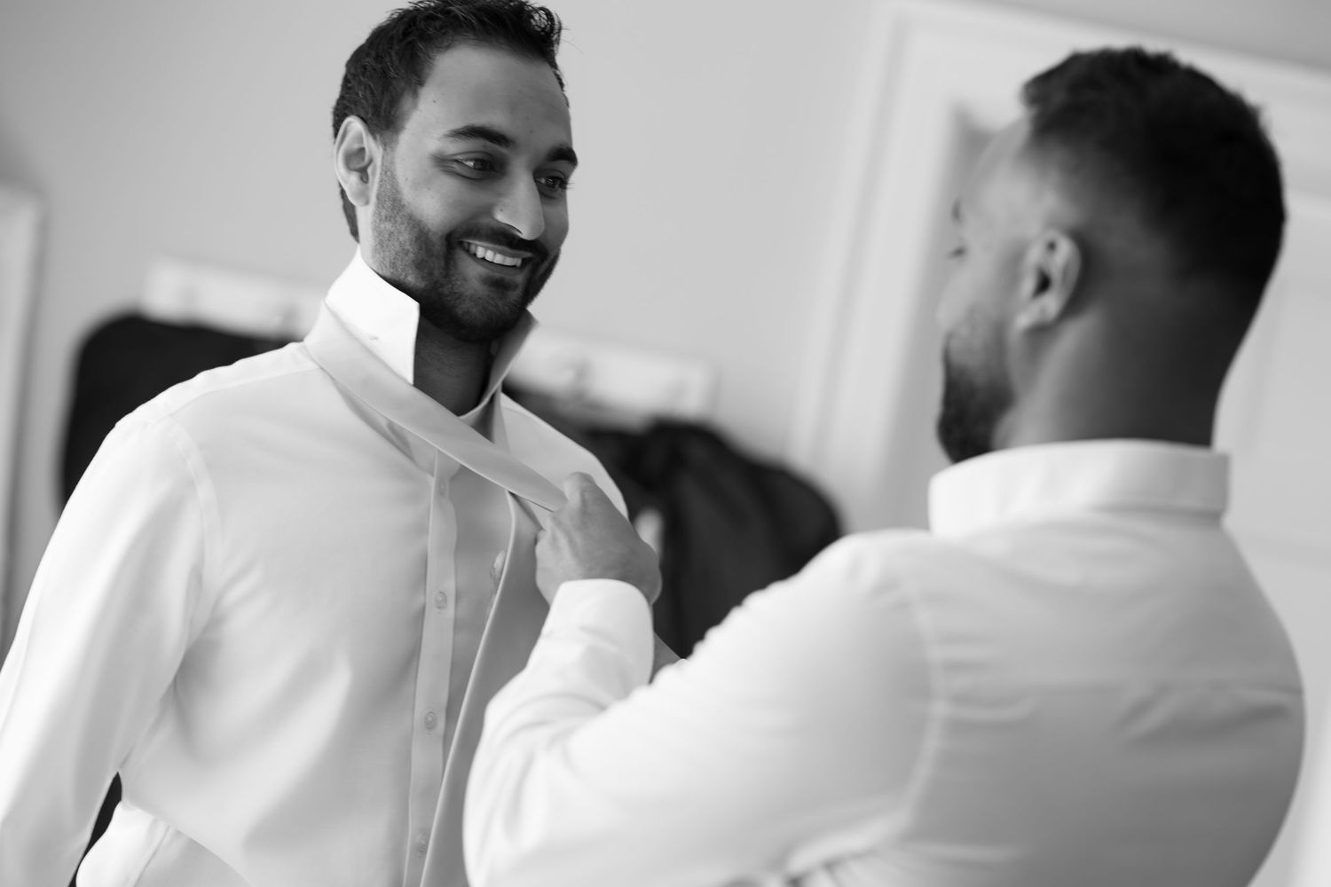 Two men in white dress shirts help each other prepare, one smiling, in a black and white wedding preparation photo.