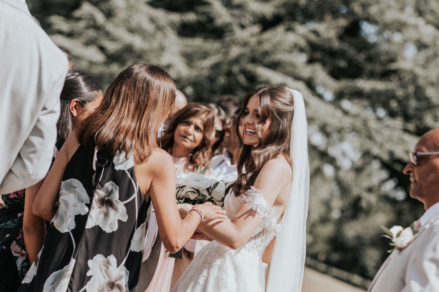 Bride in white gown smiling with guests at outdoor wedding ceremony near evergreen trees.
