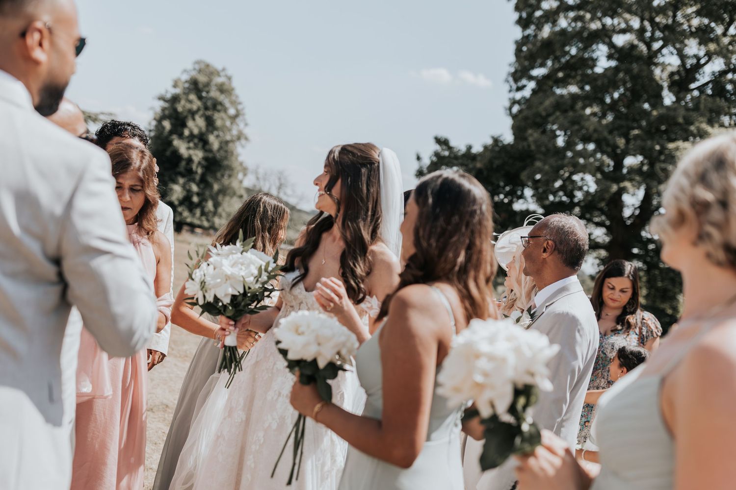 Bride in white gown and veil holds white bouquet surrounded by bridesmaids in pink dresses at outdoor wedding ceremony.