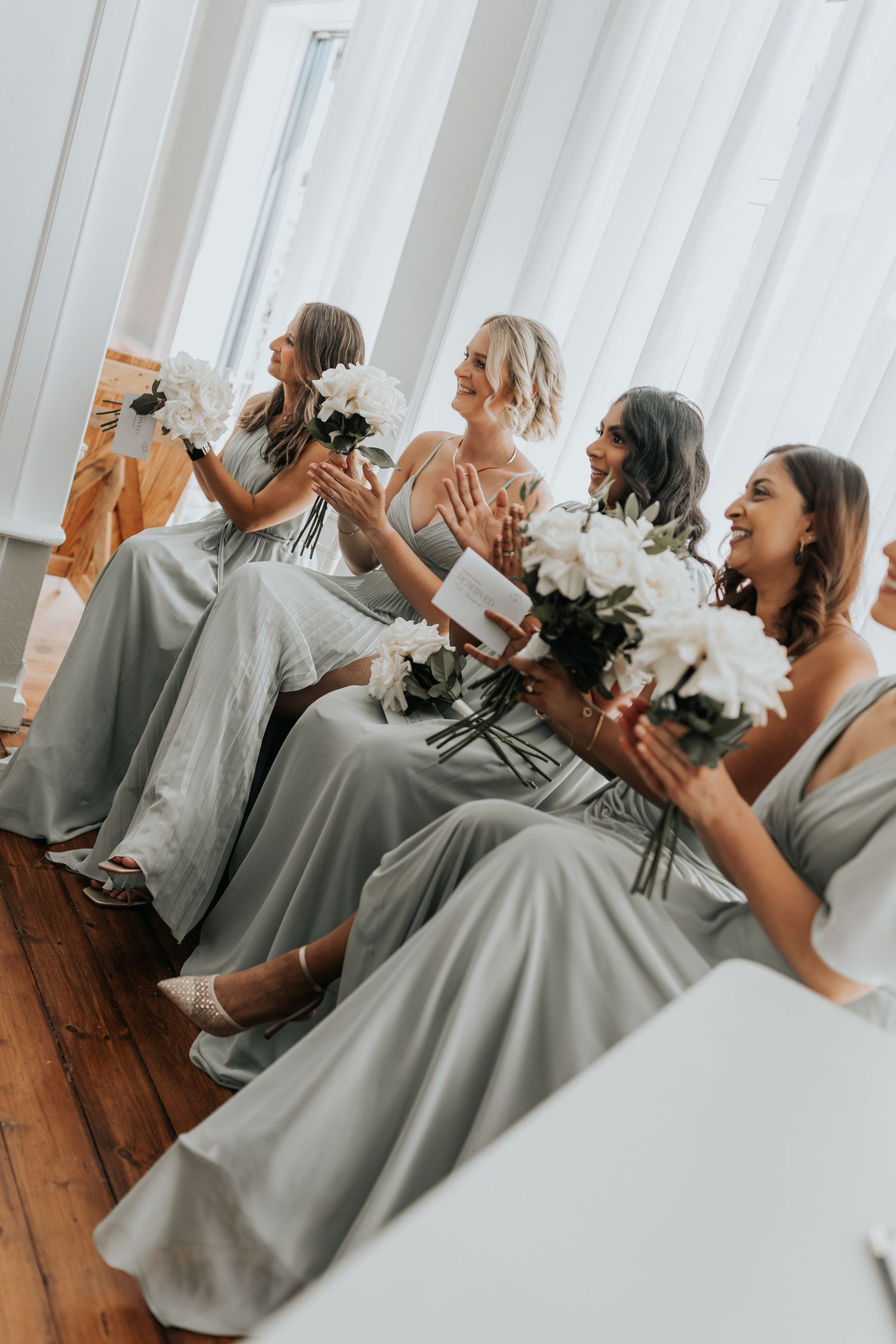 Four bridesmaids in sage green gowns holding white peony bouquets, laughing and clapping at a wedding ceremony.