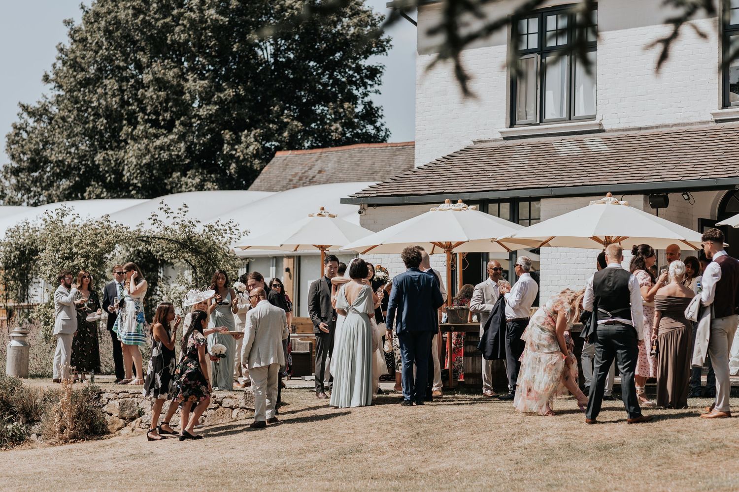 Elegant outdoor wedding reception with guests mingling on sunny patio beside white marquee and rustic country house venue.