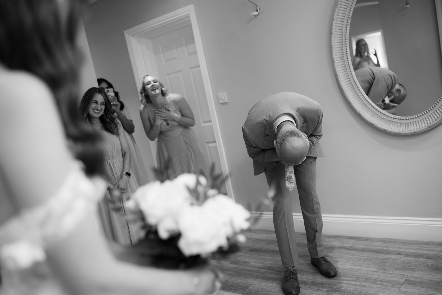 Groom bows in surprise as bridesmaids laugh seeing bride in wedding dress during getting-ready moment.