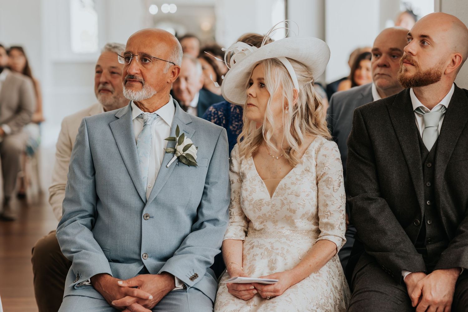 Bride in lace dress and groom in light blue suit seated during wedding ceremony with guests in background.