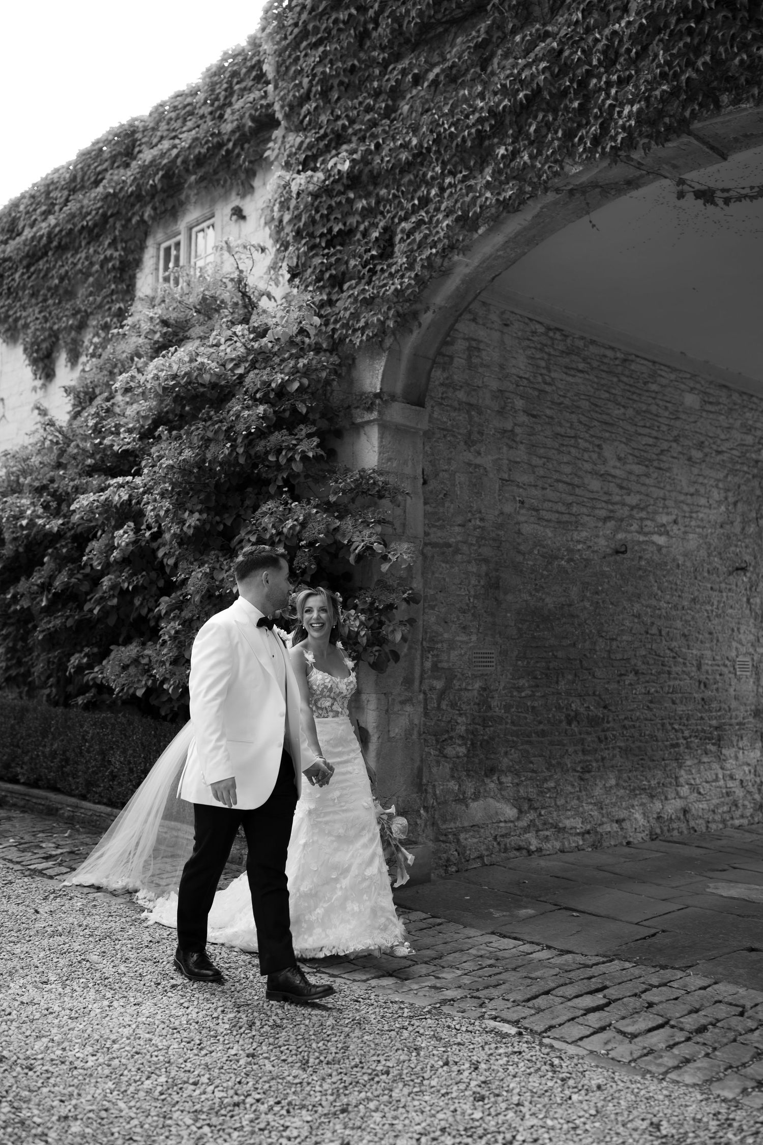 Black and white photo of a bride and groom walking hand in hand beside a stone arch covered in ivy.