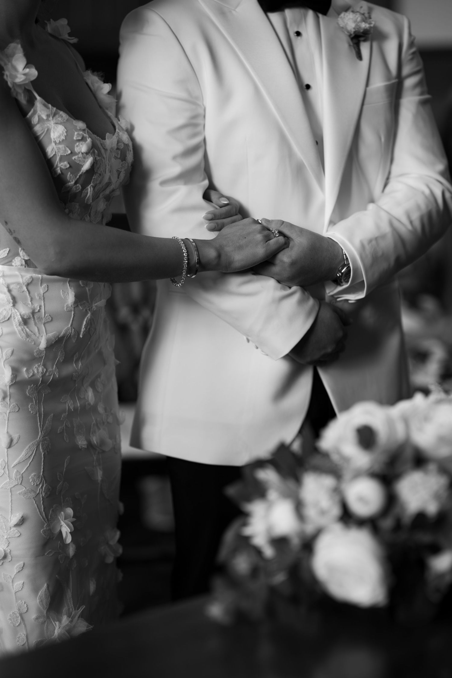 Black and white photo of bride and groom holding hands at wedding ceremony with floral arrangement.